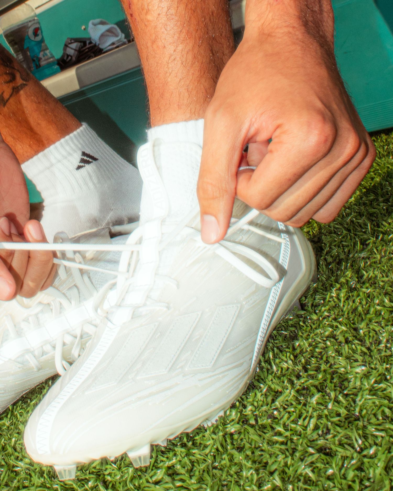 Person tying white Adidas cleats on artificial grass turf, wearing white athletic socks.
