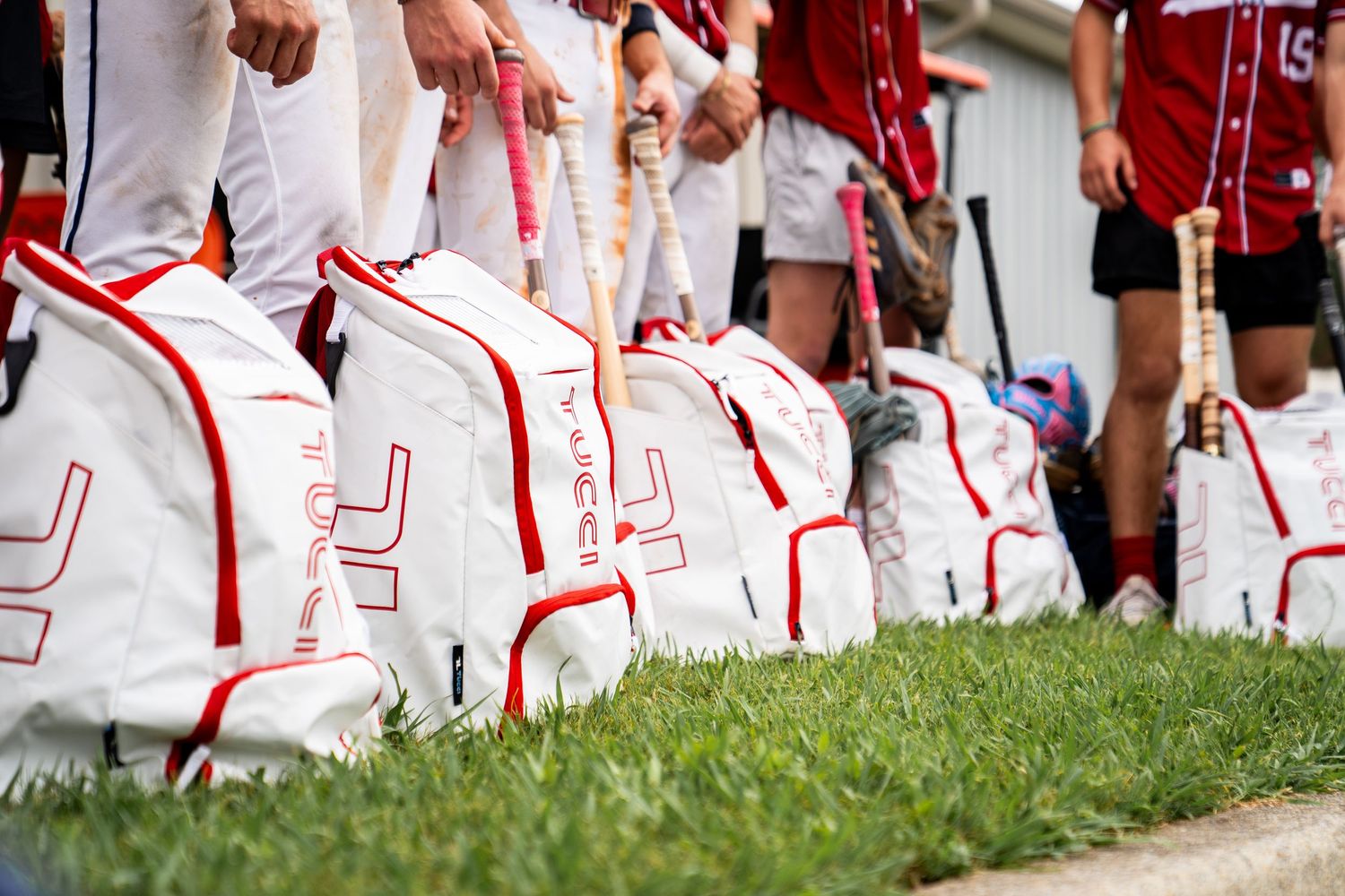 Baseball players stand beside white and red dugout bags filled with bats on a grassy field.