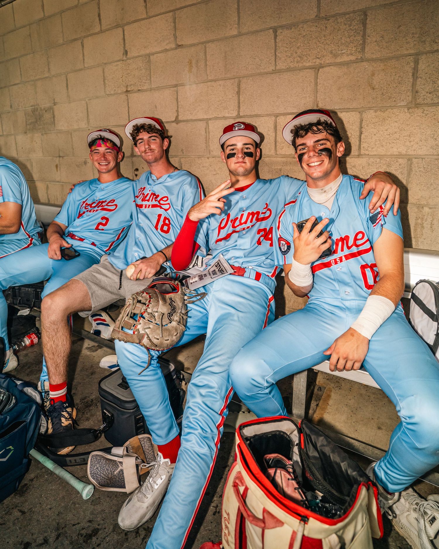 Four baseball players in blue Prime uniforms relaxing in dugout together.