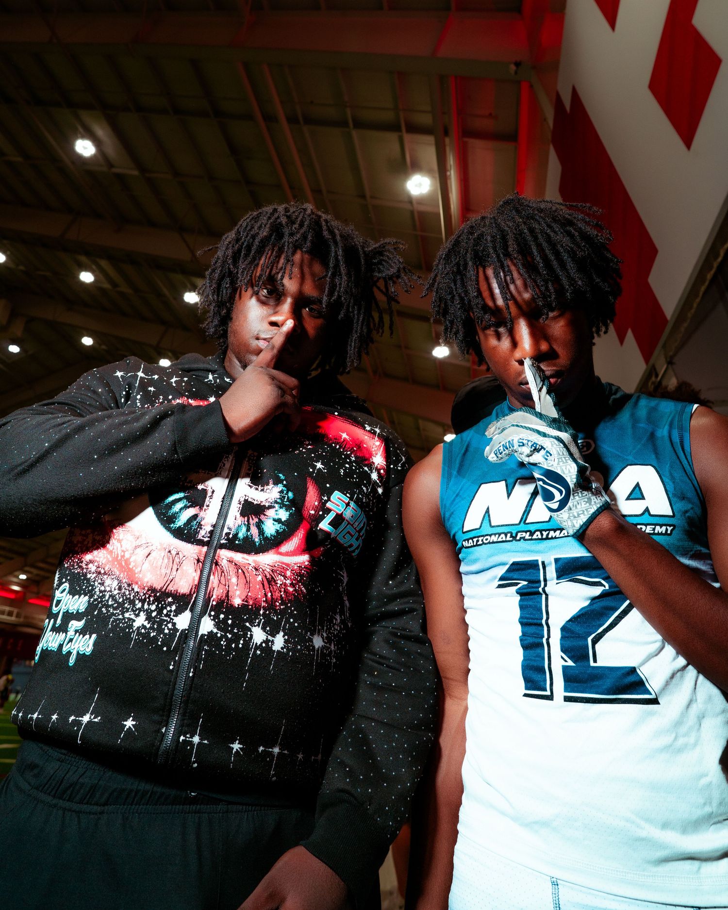 Two young men with dreadlocks pose wearing graphic streetwear, one in a dark hoodie and one in a teal NBA jersey.