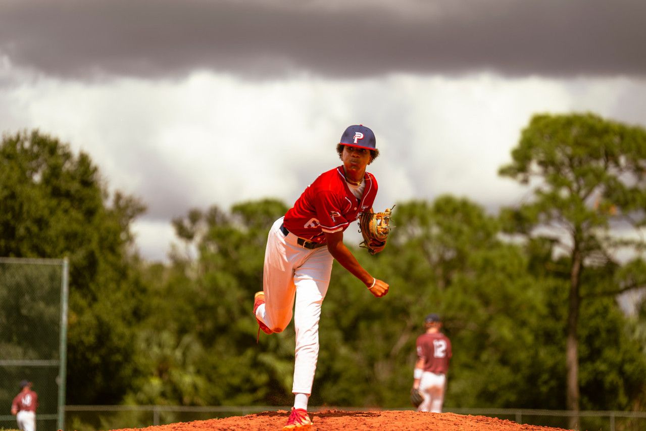 Baseball pitcher in red uniform mid-throw on mound under dramatic cloudy sky.
