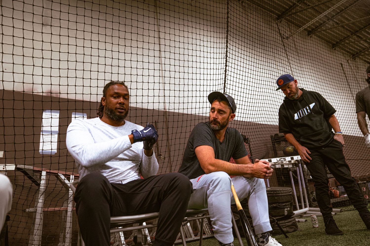 Three men sitting and standing in an indoor batting cage facility, casually conversing in athletic wear.