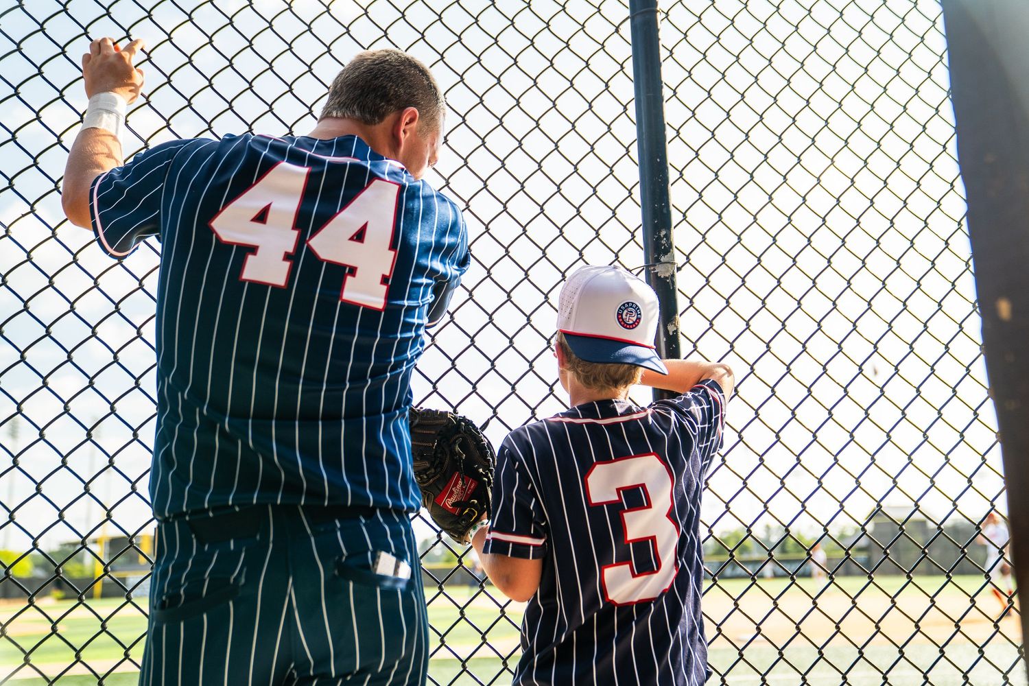 Two baseball players in navy pinstripe uniforms, numbers 44 and 3, watching a game through a chain-link fence.