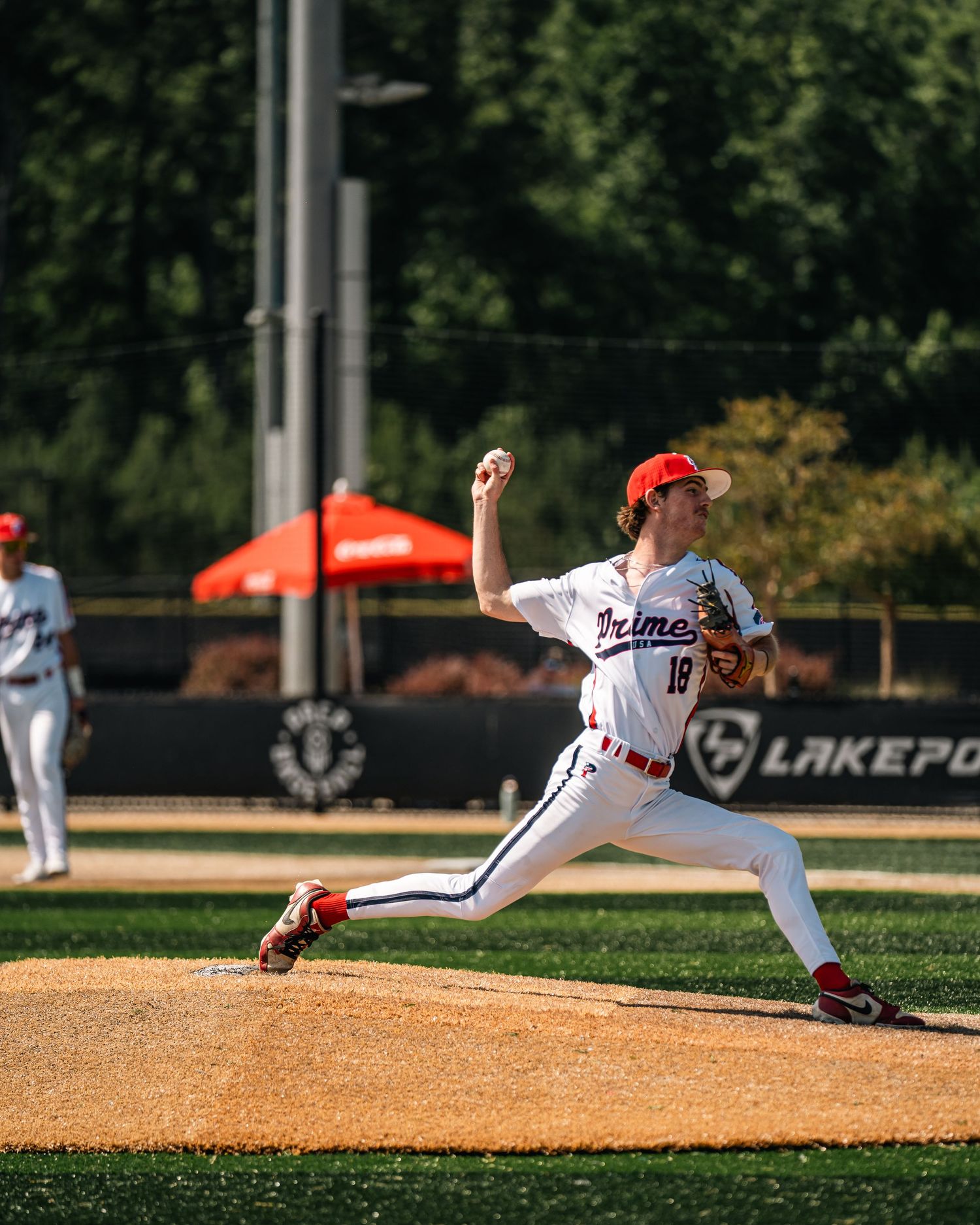 Baseball pitcher in white Primos uniform winds up to throw during a sunny outdoor game at Lakepoint.
