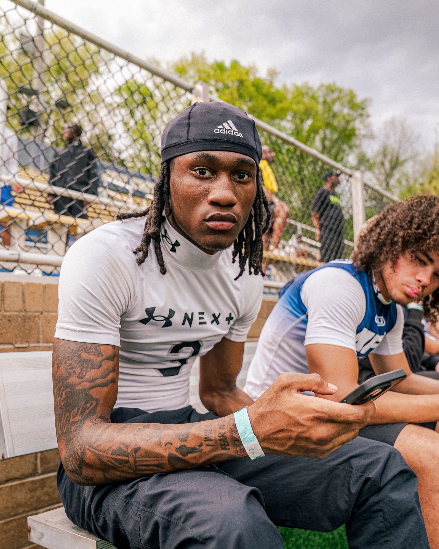 Young athlete with tattoos and Adidas cap sits in bleachers wearing Under Armour jersey at outdoor sports event.
