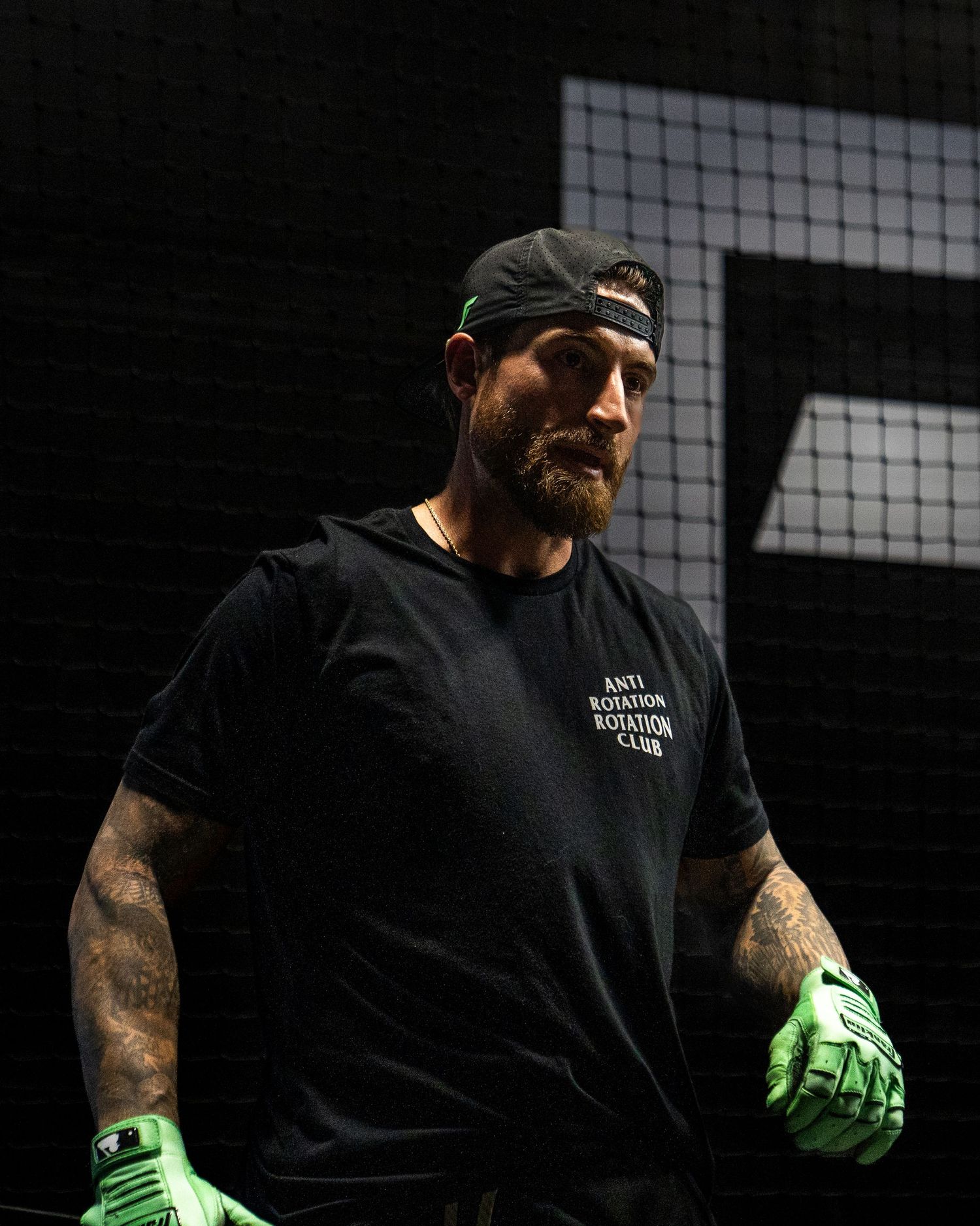 Bearded tattooed man in black Anti Rotation Club tee, cap, and green gloves stands in dark training facility.