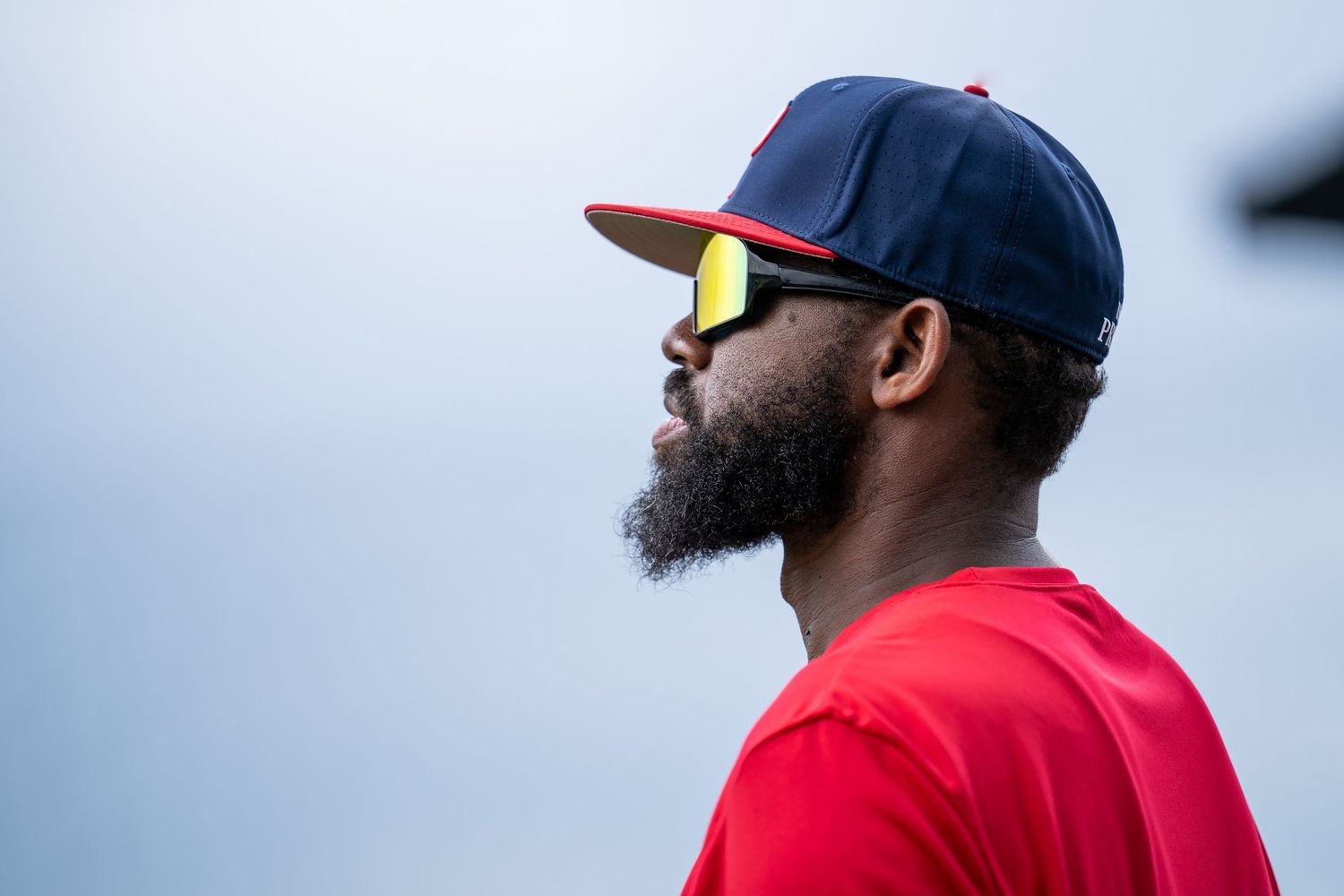 A Black man wearing a navy baseball cap, yellow-lens sunglasses, and a red t-shirt gazes upward against a cloudy sky.
