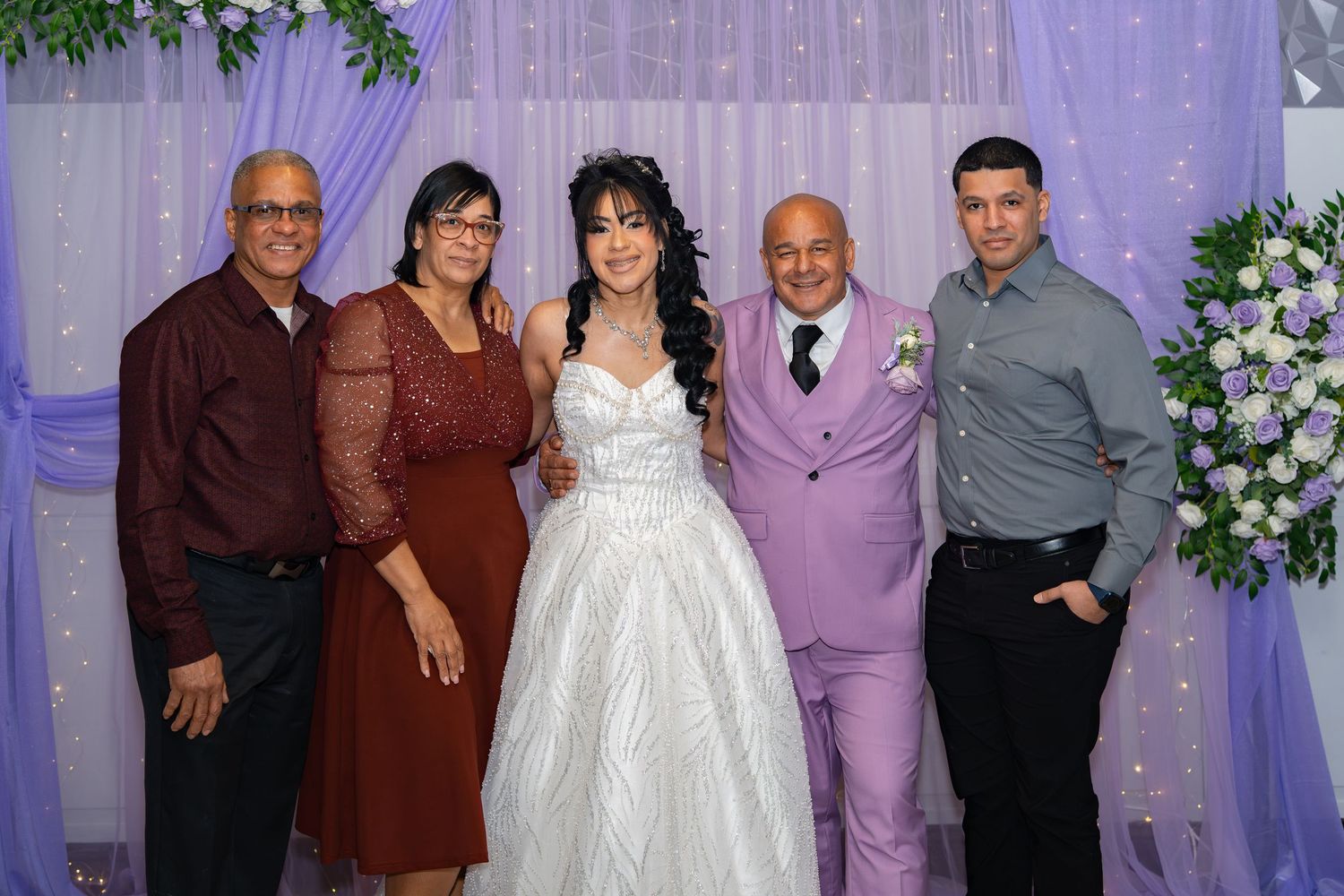 Wedding party of five poses together in front of a decorated backdrop with white and purple flowers.