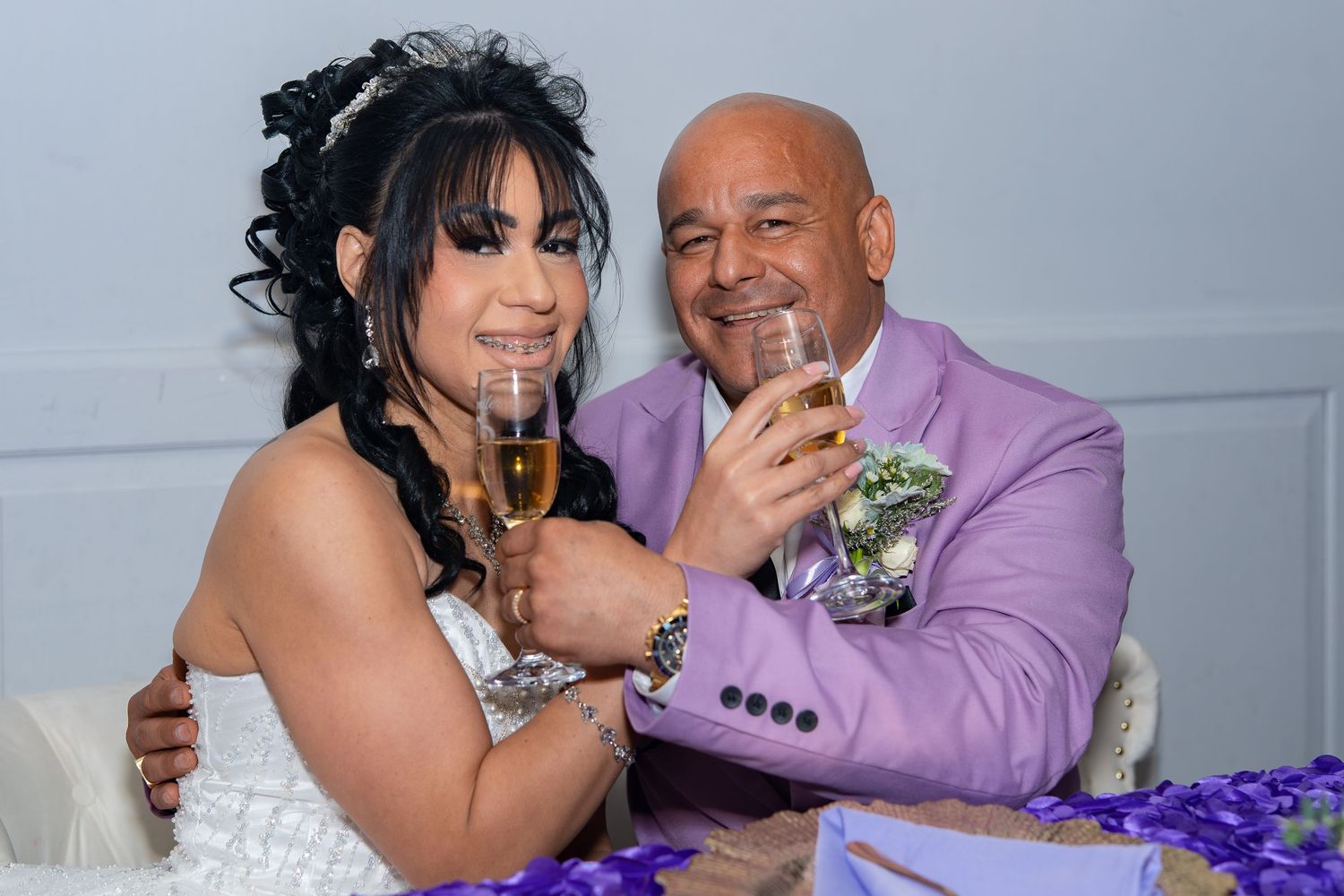 Newlyweds clink champagne glasses together smiling at their wedding reception table.