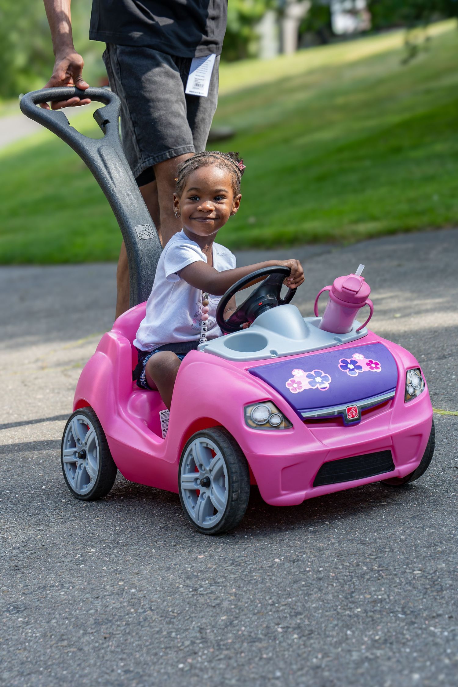 Smiling toddler girl riding a pink Step2 push car on a paved path outdoors.