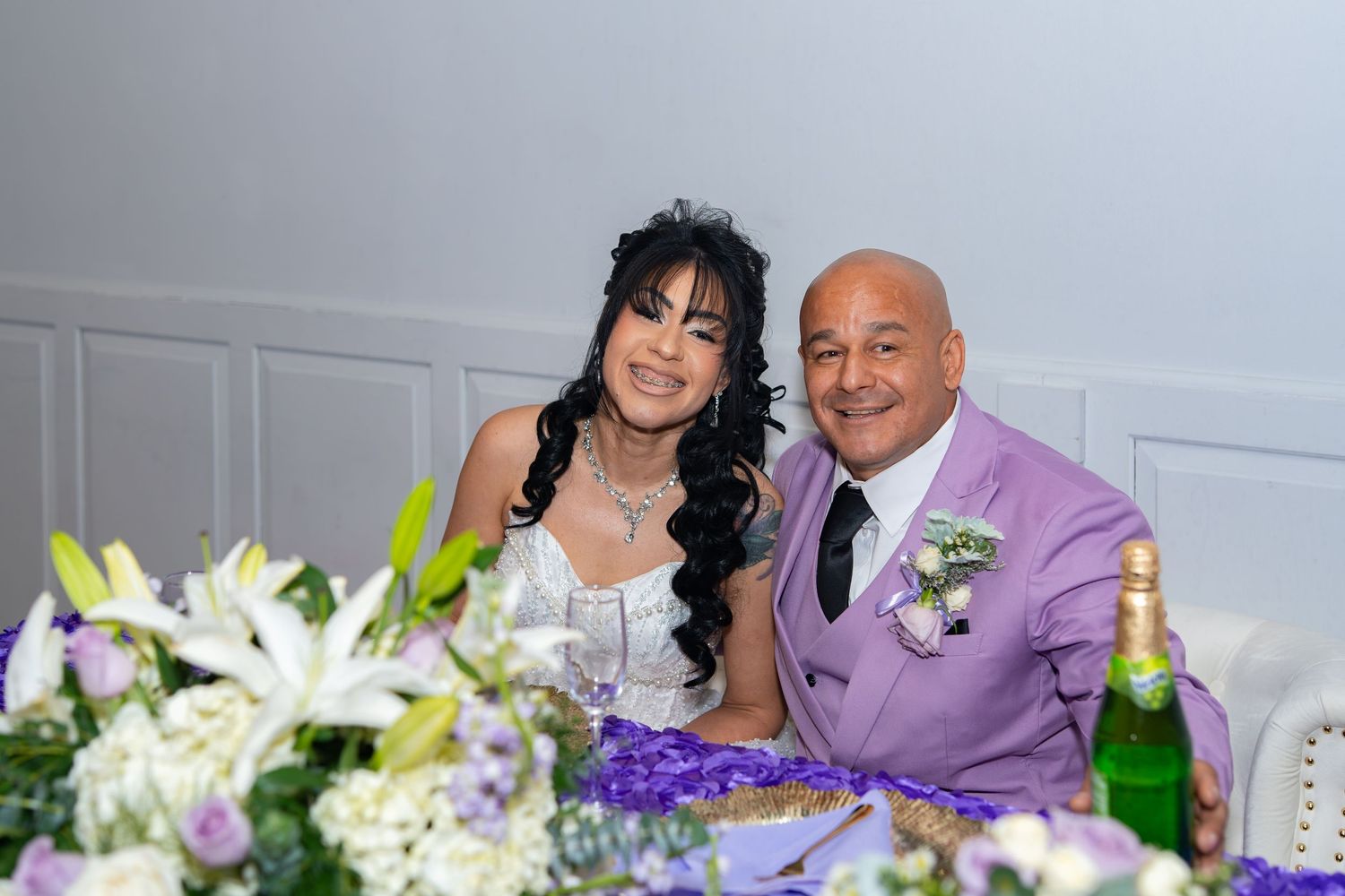 Bride and groom in lavender suit sit together at their floral-decorated wedding reception table.