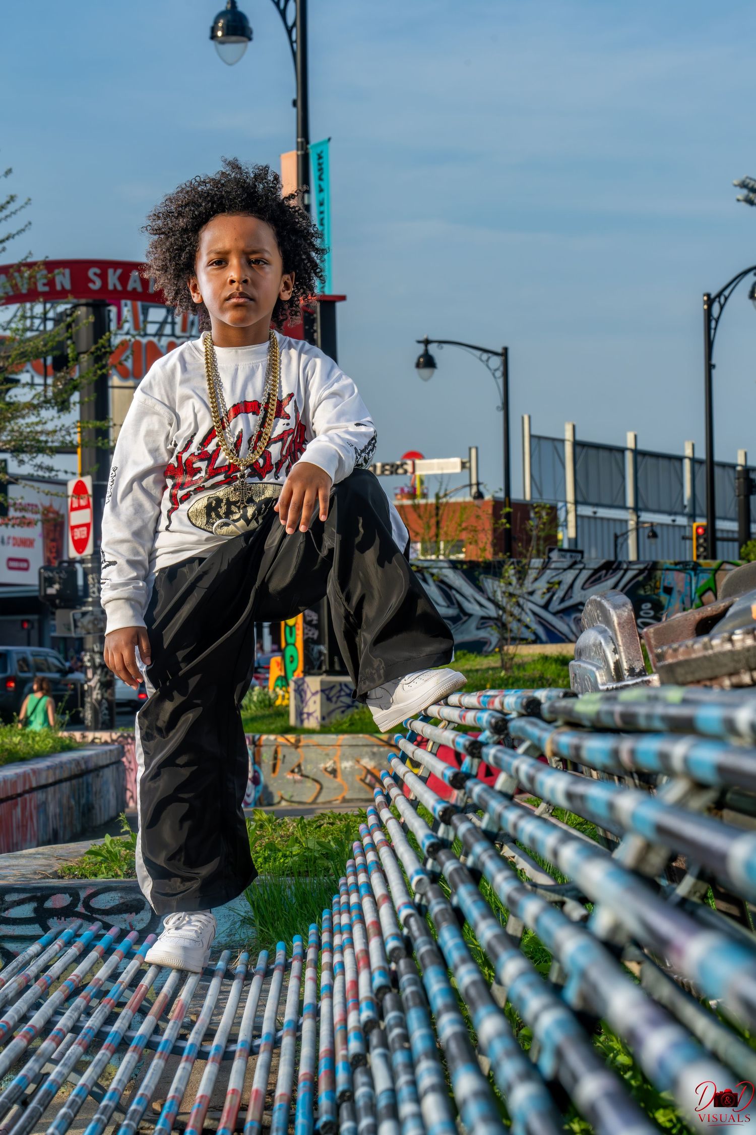 Young boy in urban streetwear poses confidently at a skate park with graffiti art in the background.