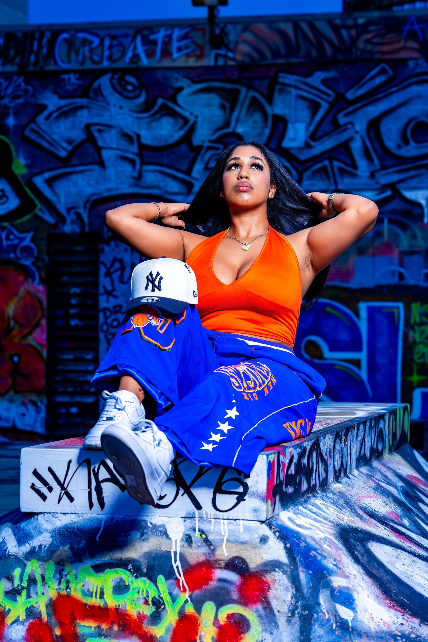 Lifestyle portrait of a woman in an orange top and blue pants seated against colorful graffiti.