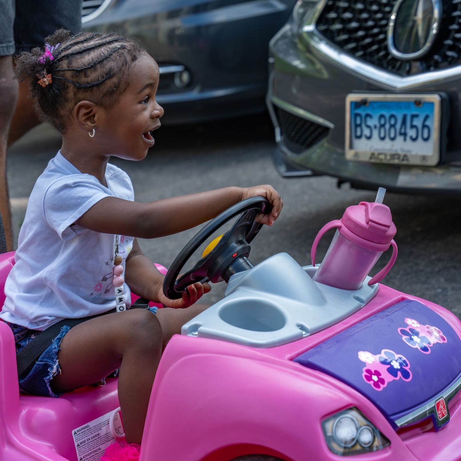 Excited young girl sitting in a pink toy car outdoors, gripping the steering wheel joyfully.