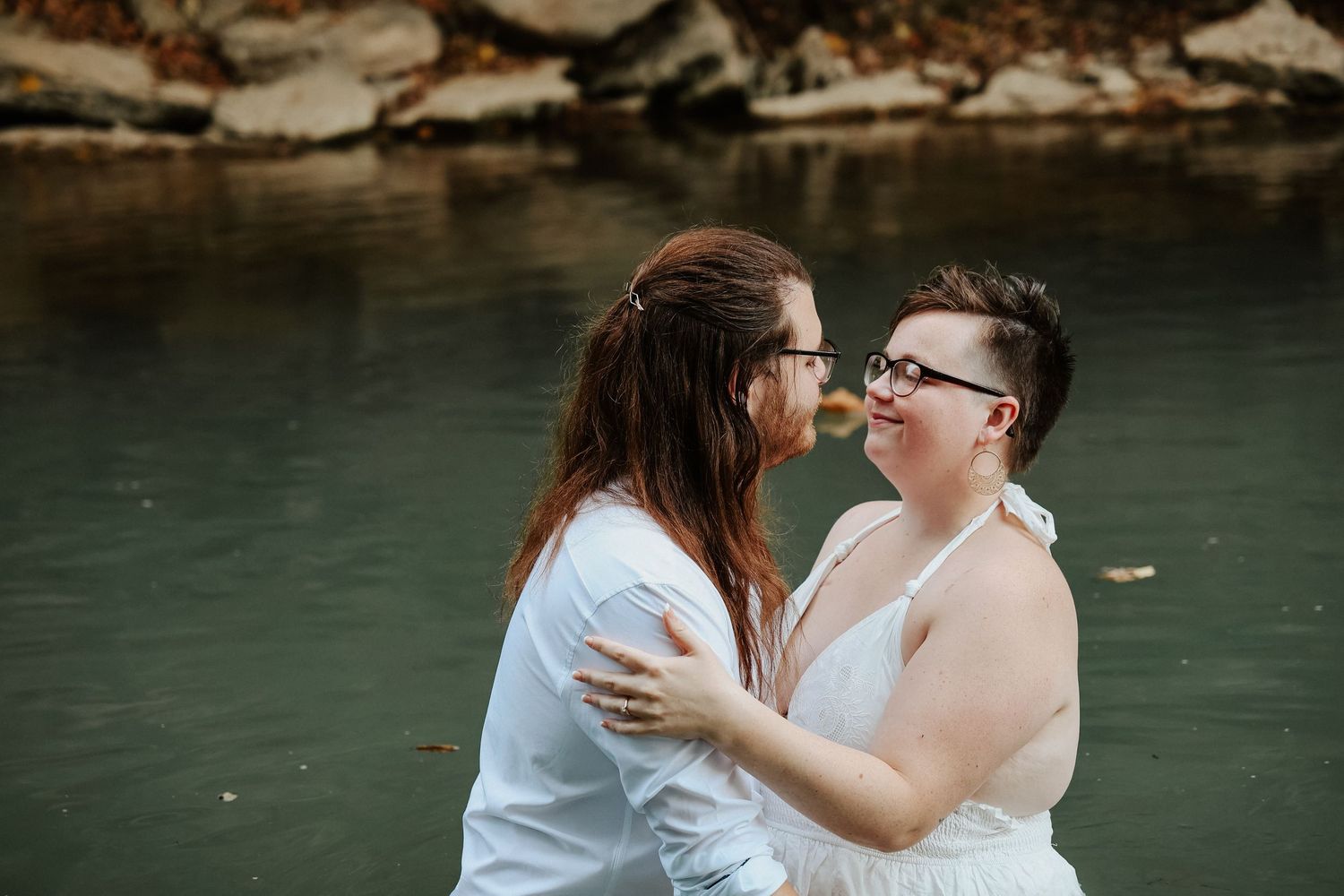 Two brides embrace and smile at each other while standing in a river during their outdoor wedding elopement.