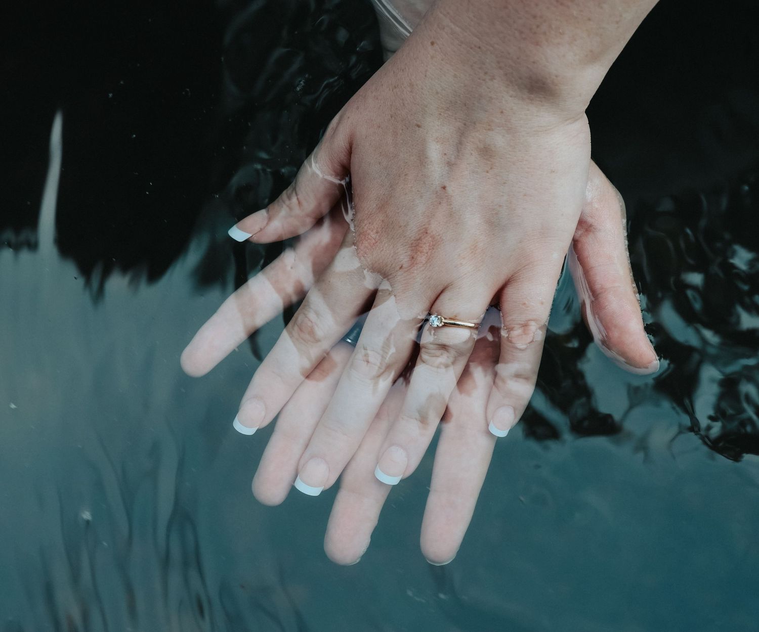 Two hands with white French tip nails and a delicate ring rest together over dark water.