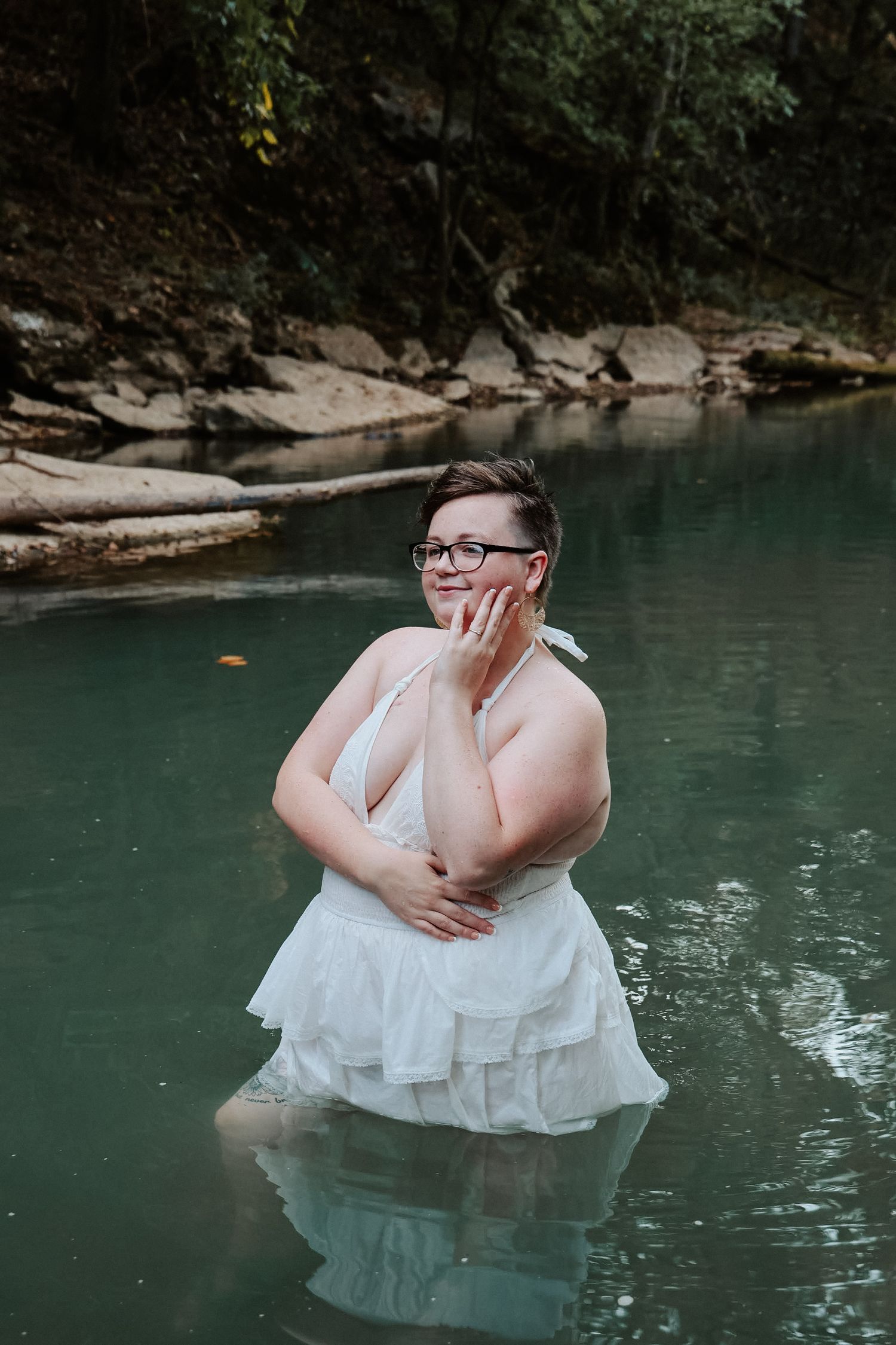 A plus-size woman in a white dress stands in a serene green river, posing thoughtfully while wearing glasses.