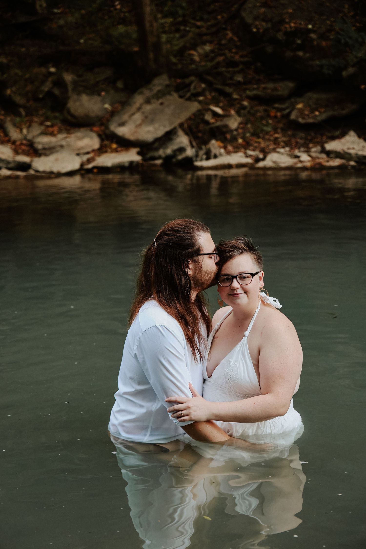 Couple embracing in a serene river, wearing white outfits, sharing a romantic moment near rocky shores.