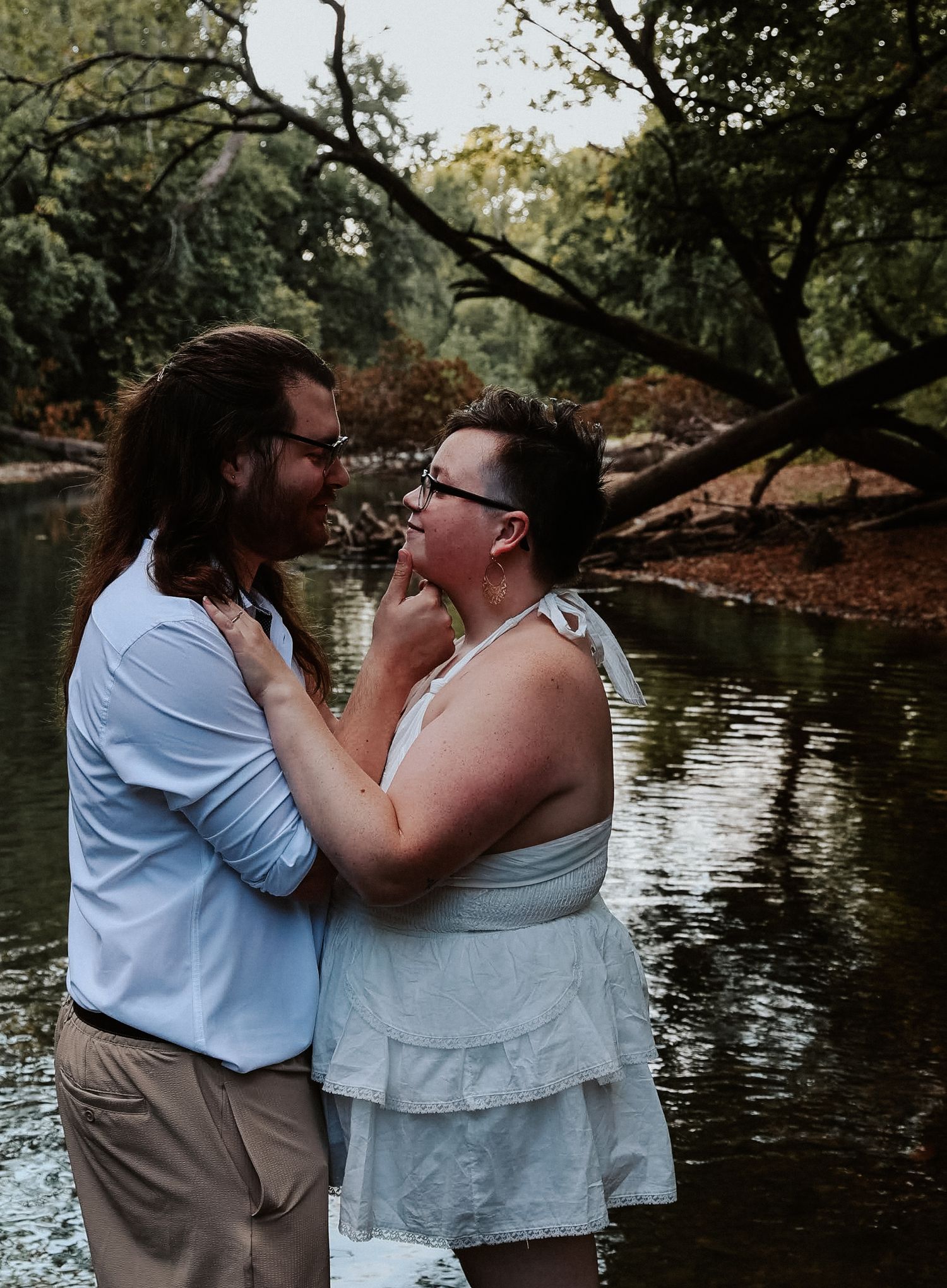 Couple embracing romantically by a serene river surrounded by lush green trees in an outdoor setting.