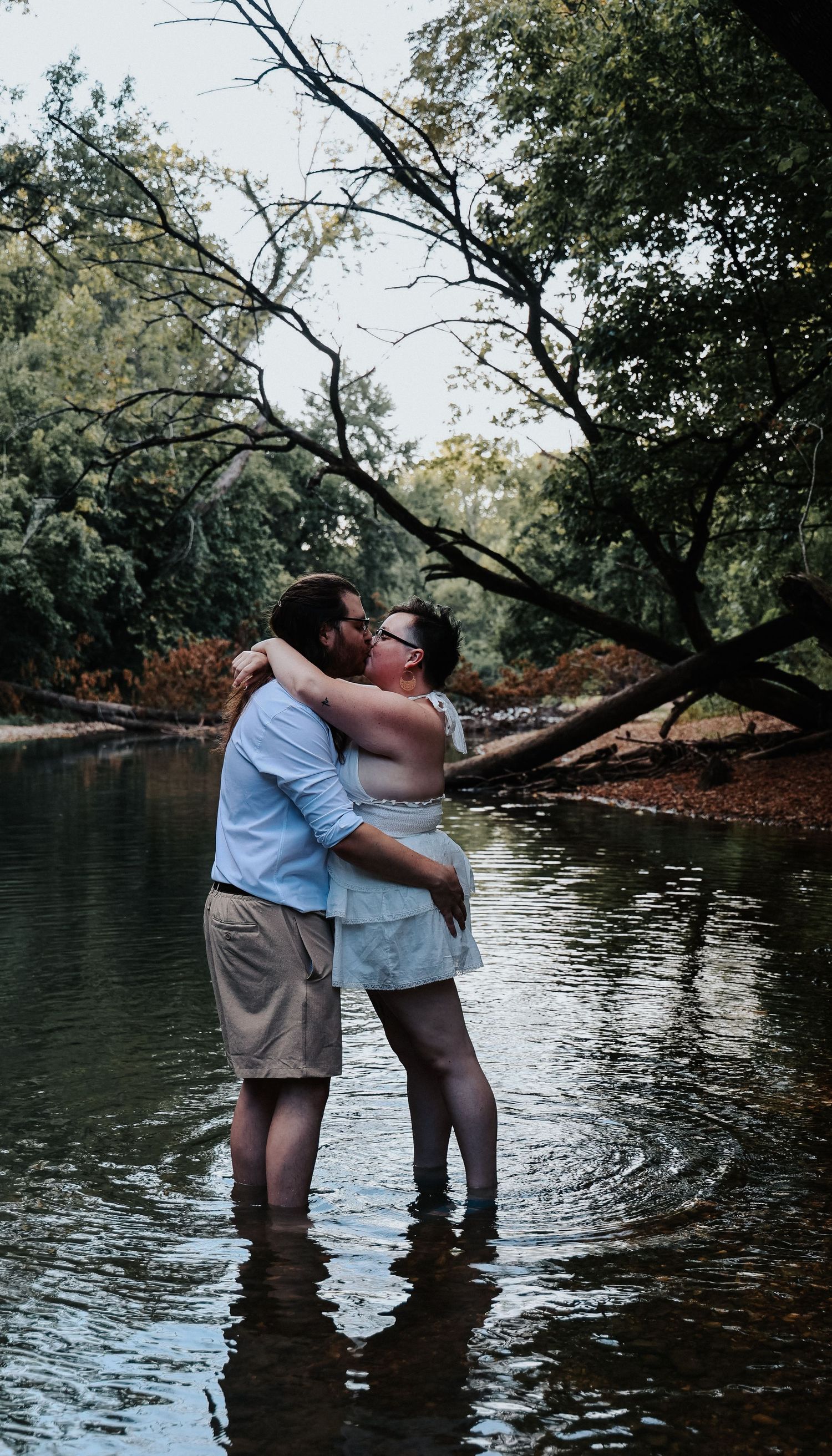 Couple embracing romantically while standing in a shallow river surrounded by lush green trees.