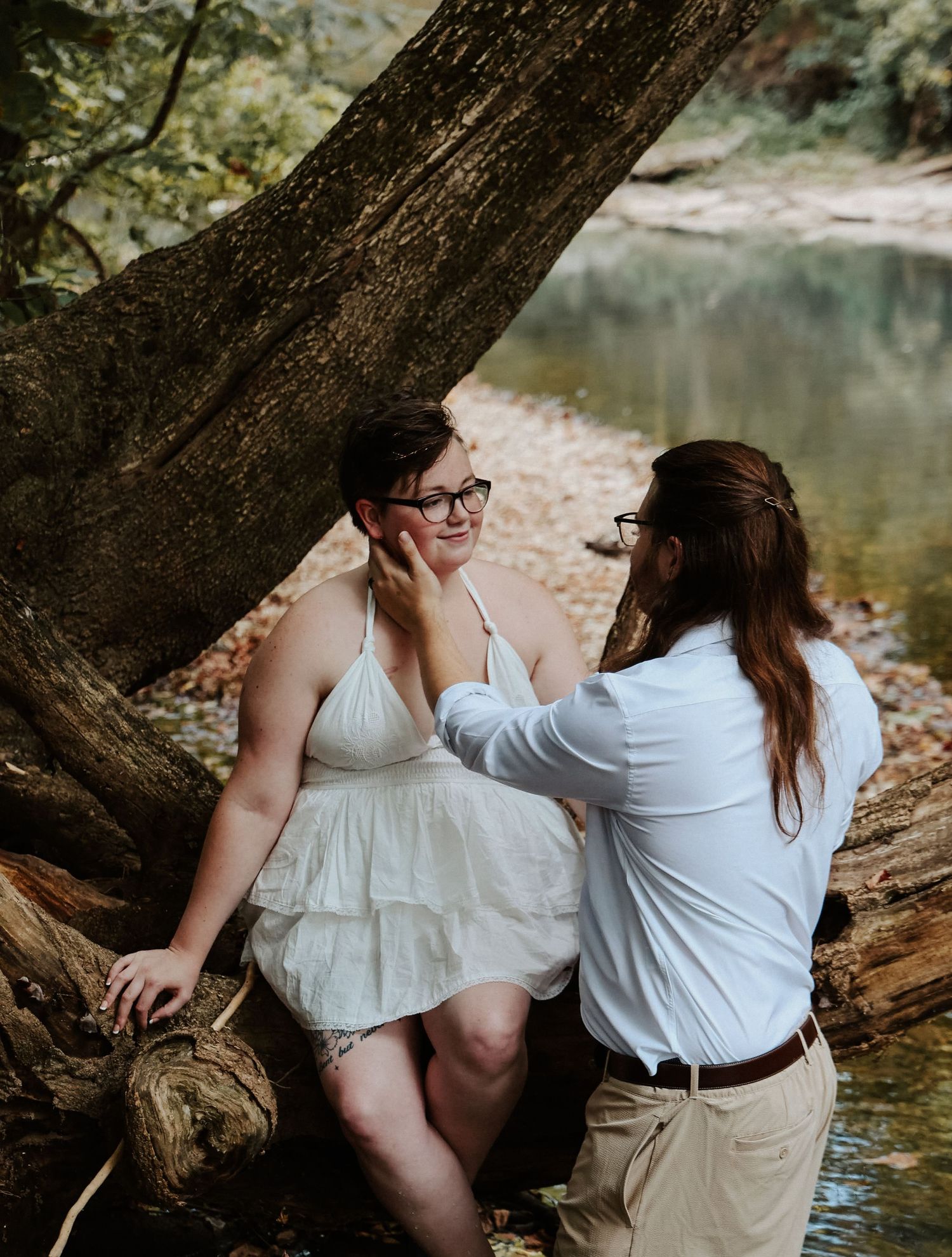 Couple shares a tender moment by a creek, one in a white dress, the other in a white shirt, surrounded by nature.