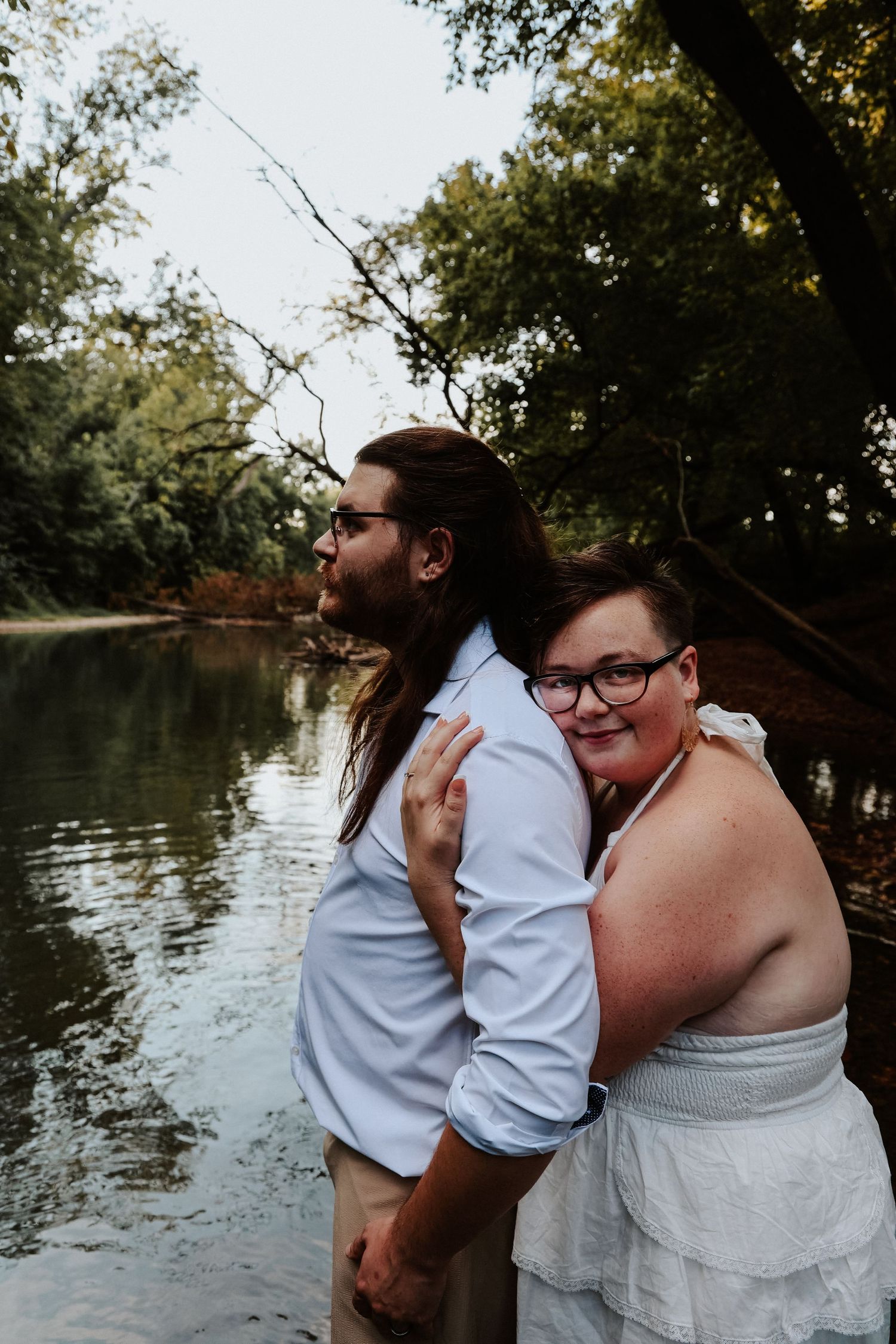 Couple embracing by a serene river surrounded by lush green trees, woman in white dress hugging man in blue shirt.