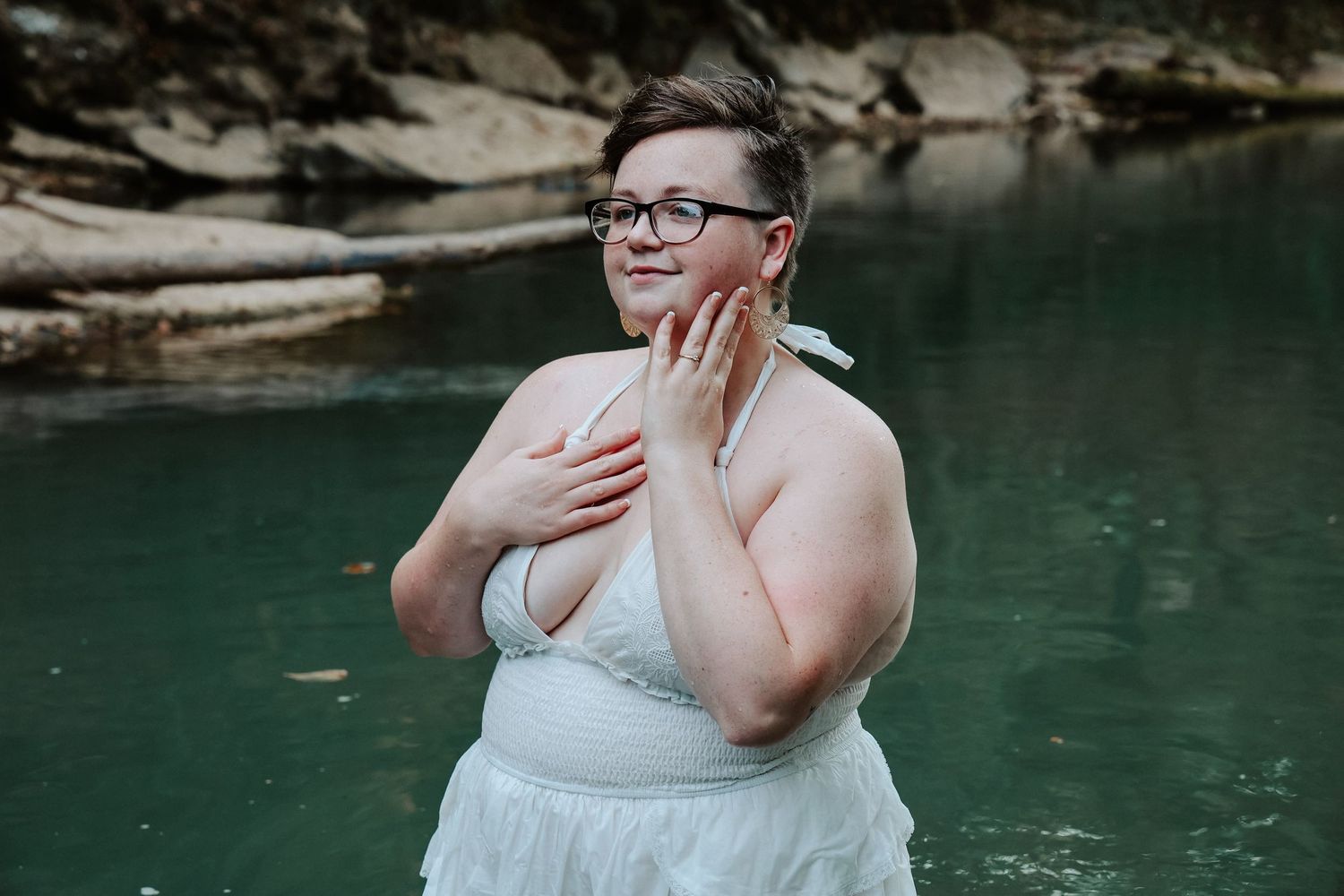 Person in white dress standing in natural pool with rocky background, hands raised gracefully near face.