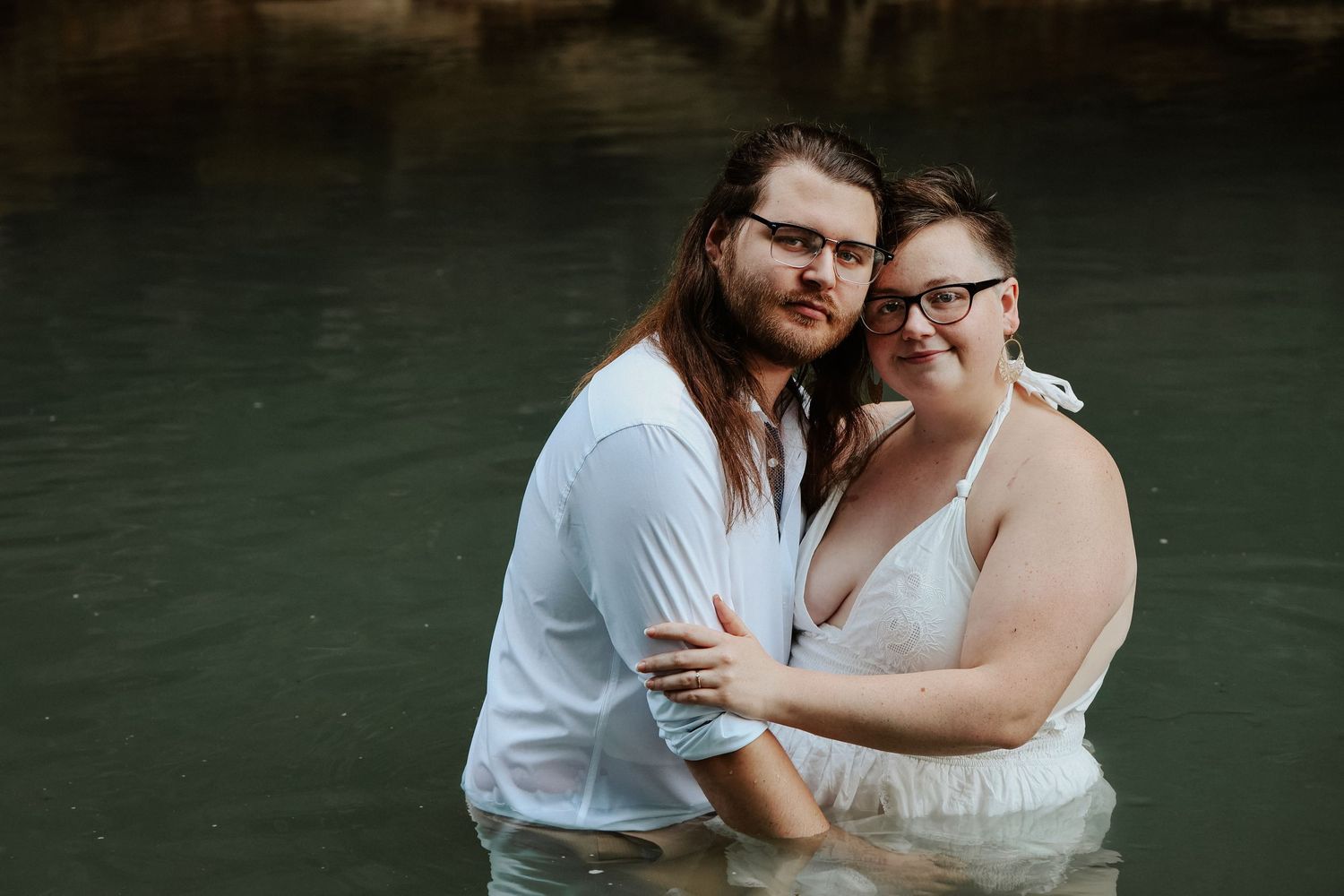 Couple embracing in water wearing white outfits, romantic outdoor engagement photo session in a natural setting.