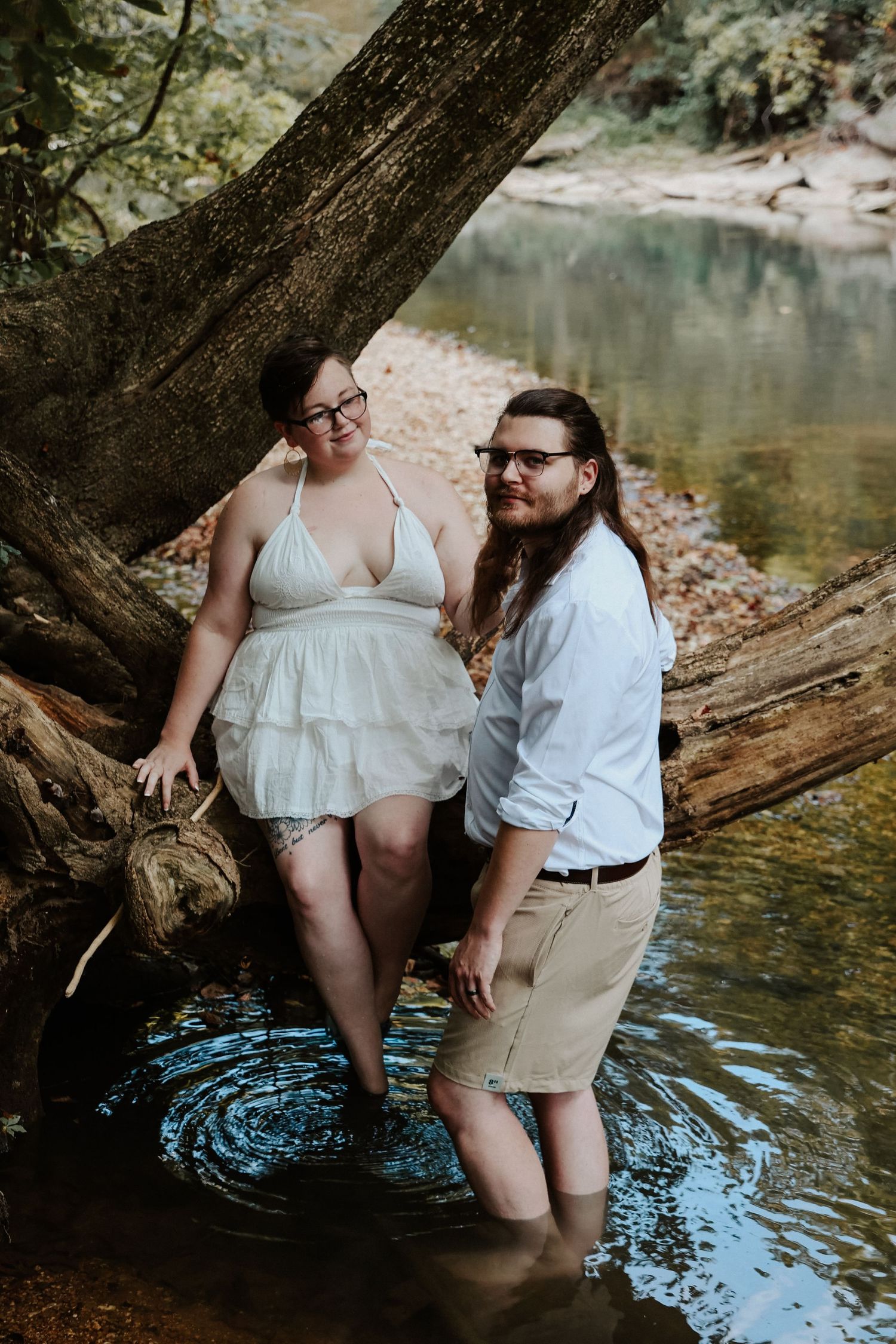 Couple in white attire standing in a shallow creek surrounded by trees during an outdoor engagement session.