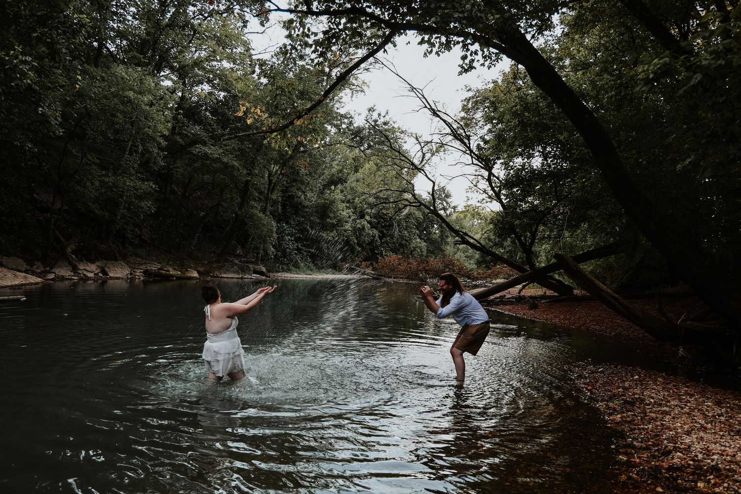 Two people playfully wade in a serene forest creek, surrounded by lush green trees in a candid outdoor moment.