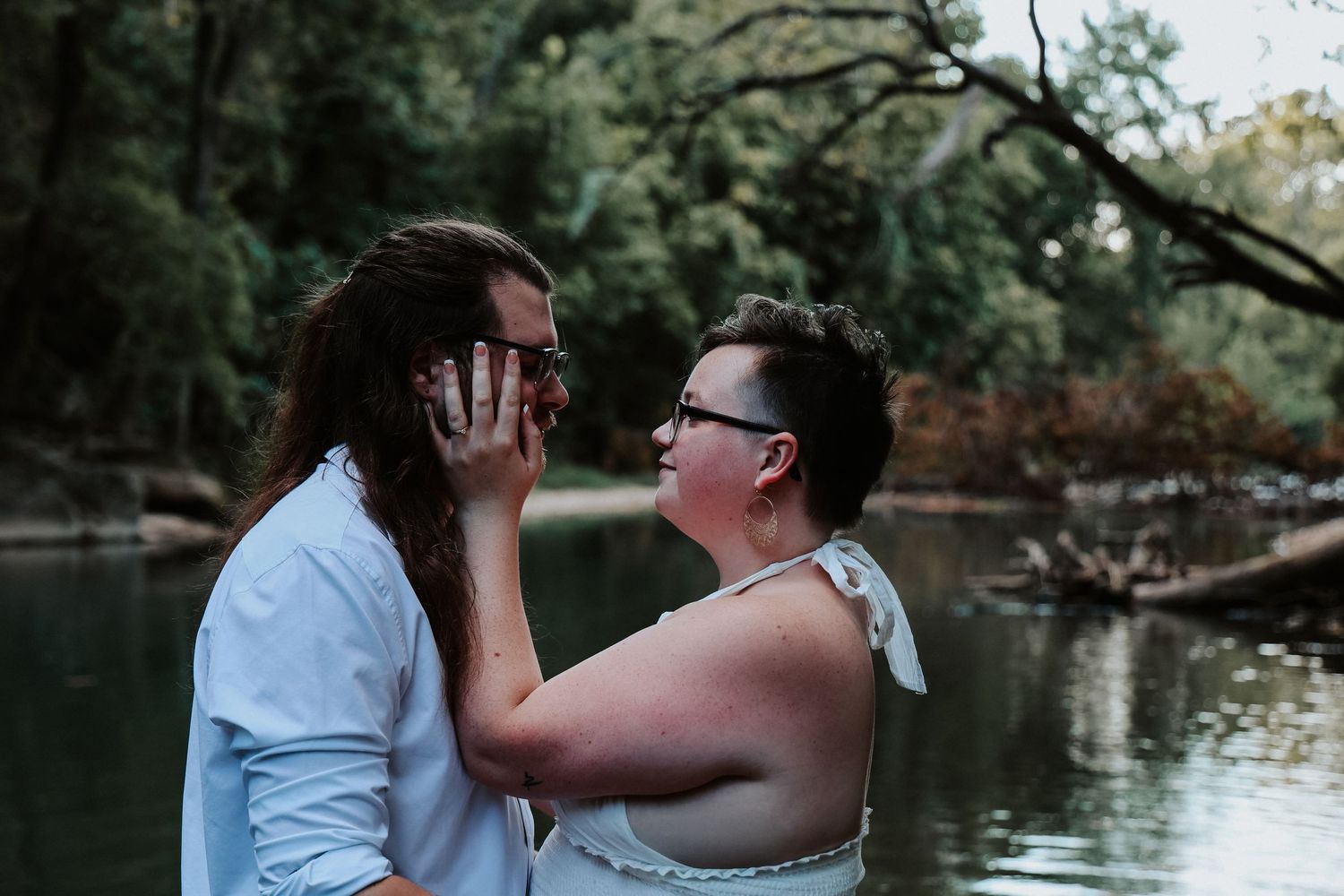 Couple shares a romantic moment by a serene river surrounded by lush green trees, in an outdoor wedding photo.