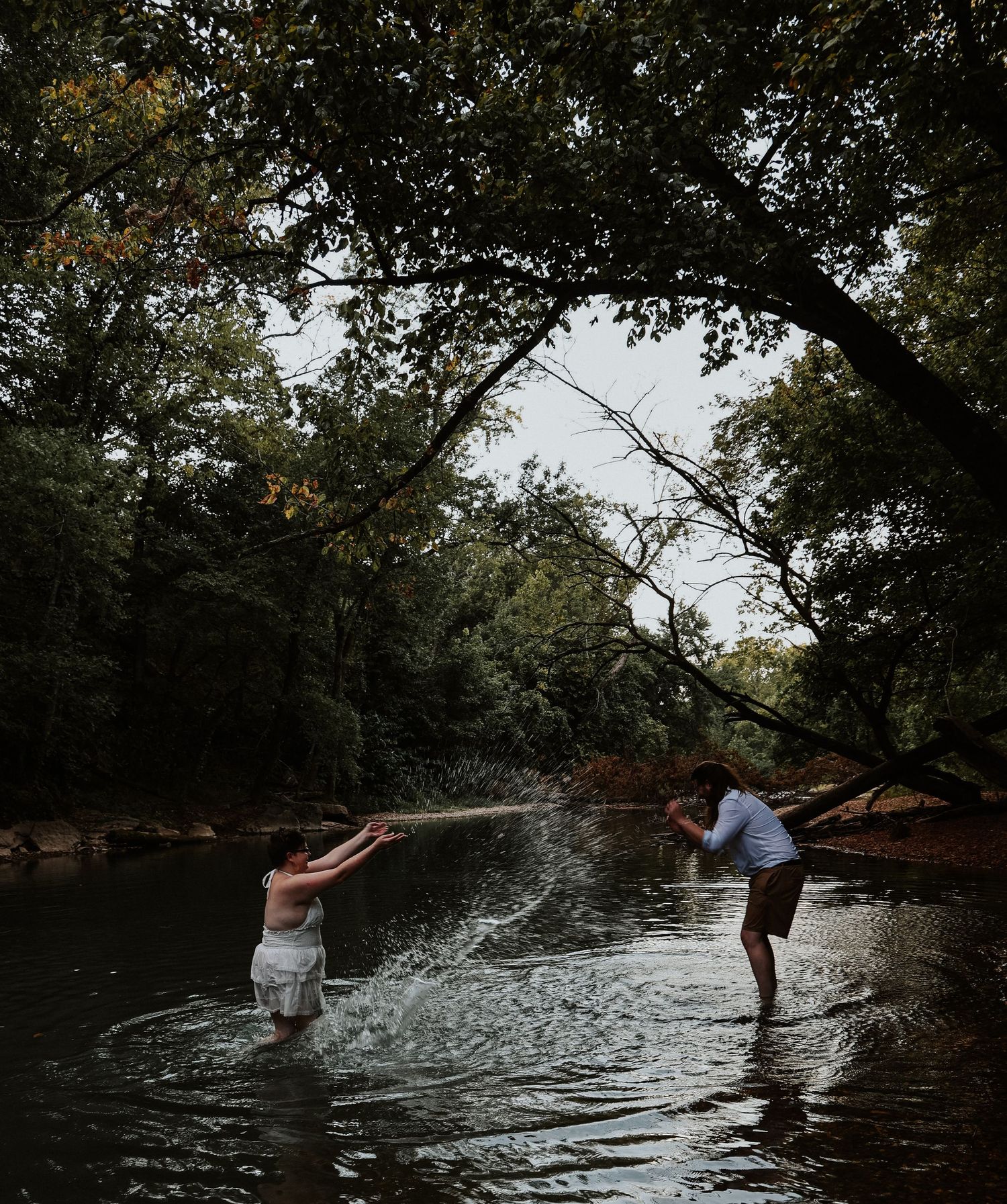 Couple playfully splashing water in a serene forest creek surrounded by lush green trees.