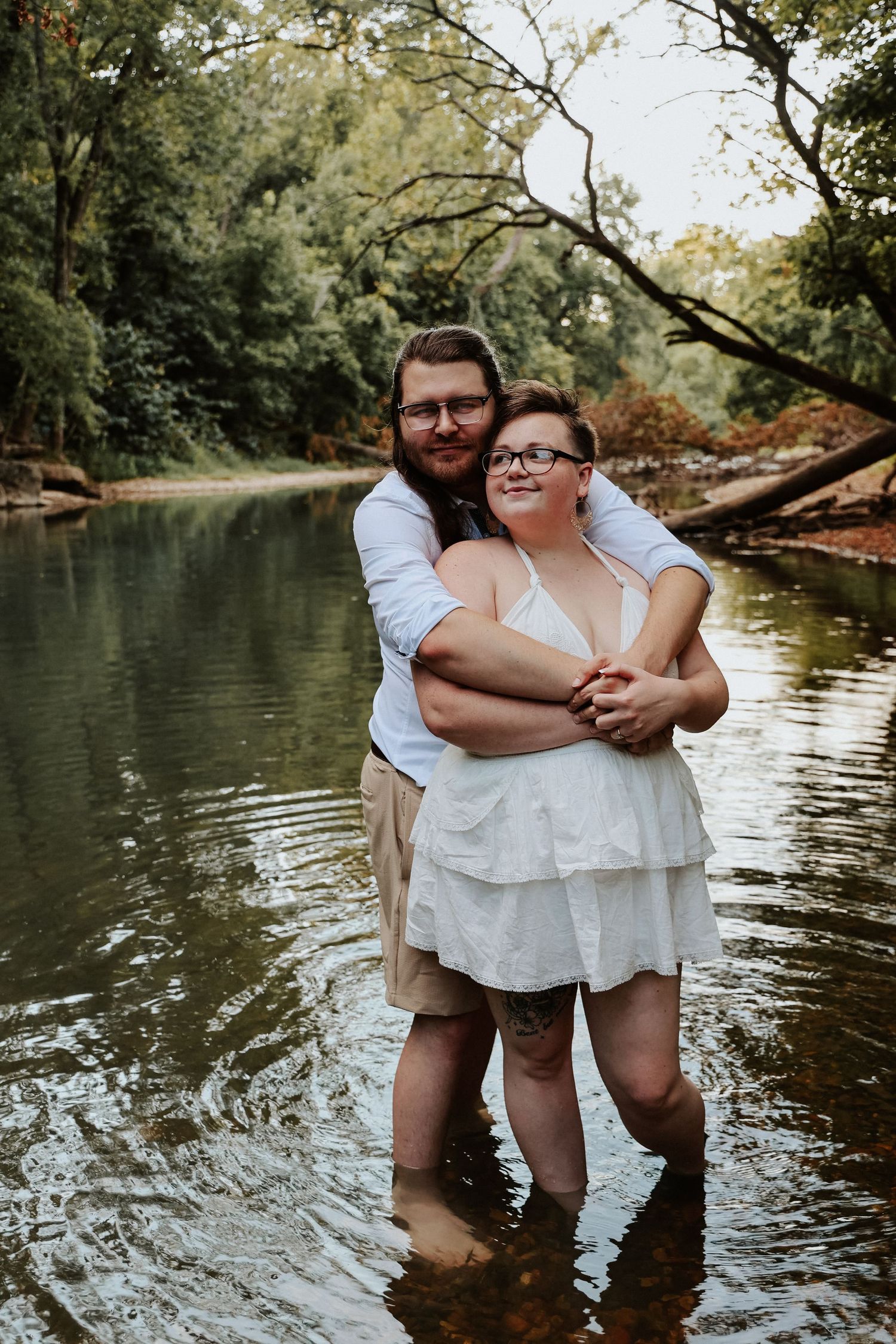 Couple embracing in a shallow creek surrounded by lush green trees, wearing white and khaki outfits.