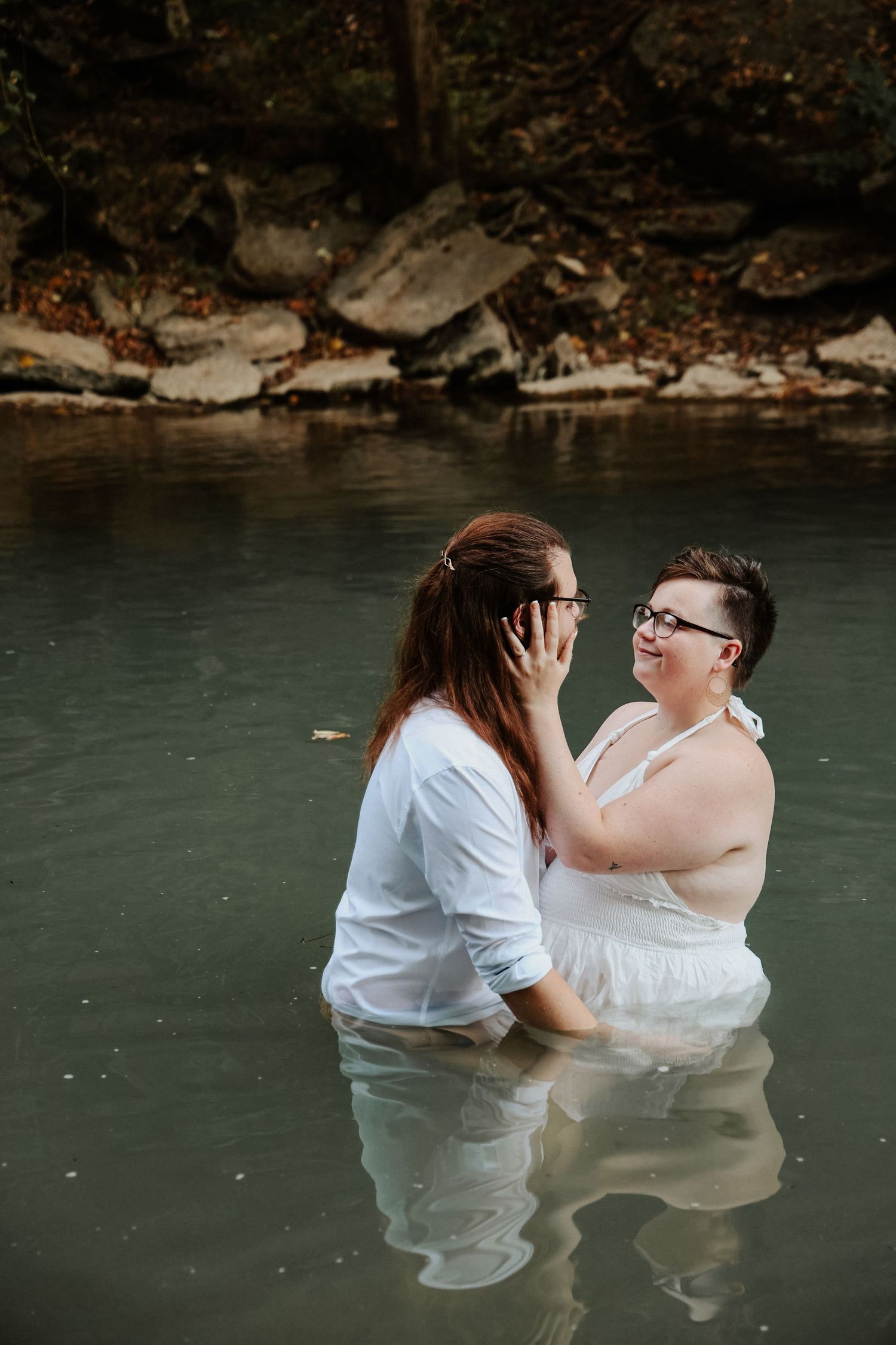 Two women in white dresses share a tender moment standing together in a serene river surrounded by rocks.