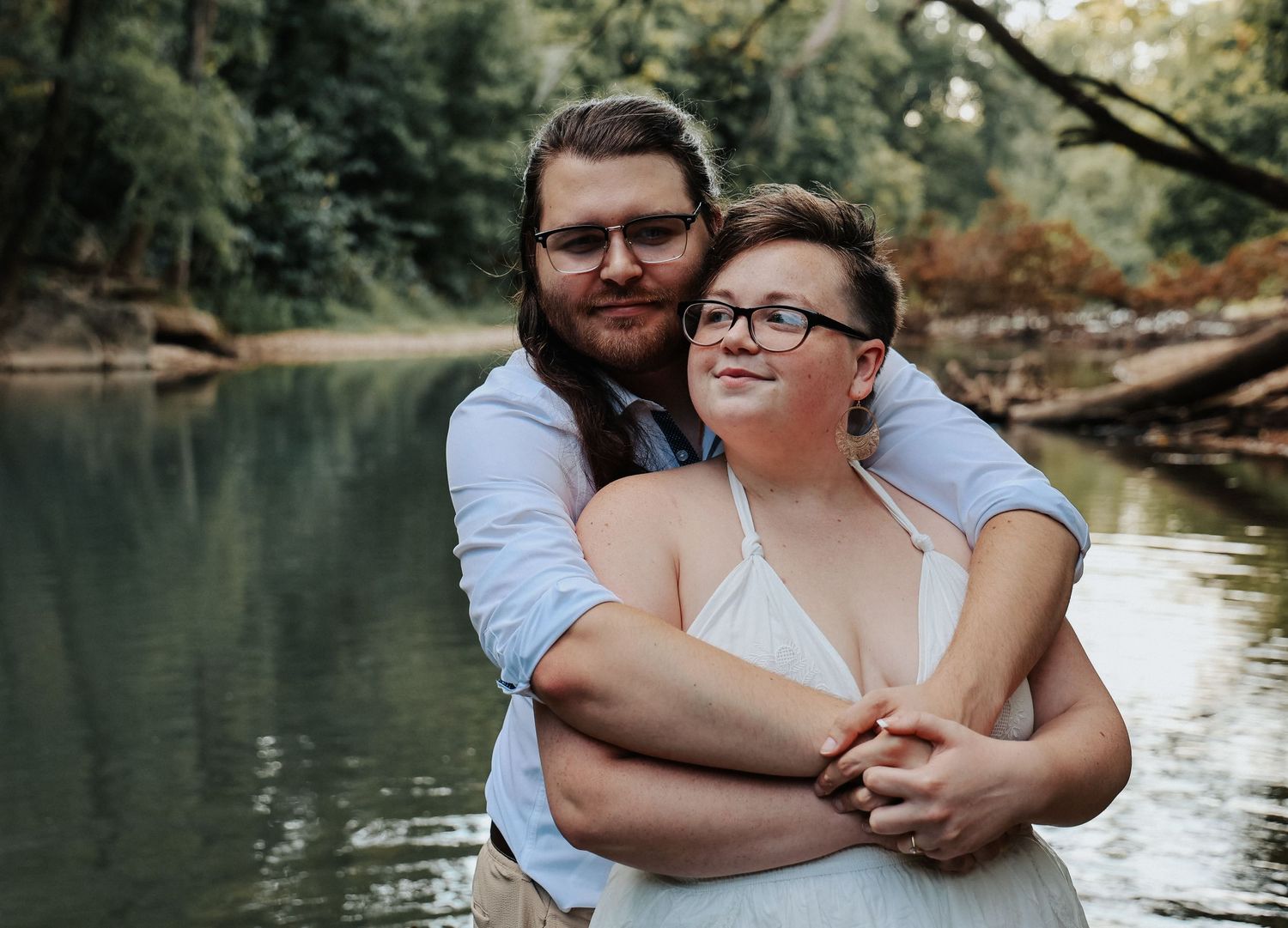 Couple embracing near a serene forest river, woman in white dress with glasses, man in blue shirt.