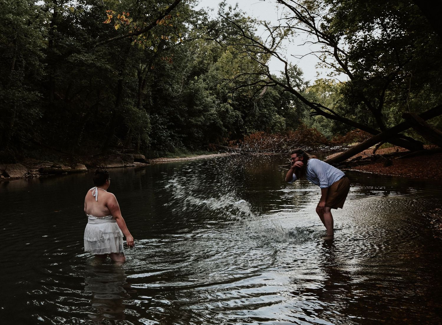 Two people wading in a calm forest river surrounded by lush green trees on an overcast day.