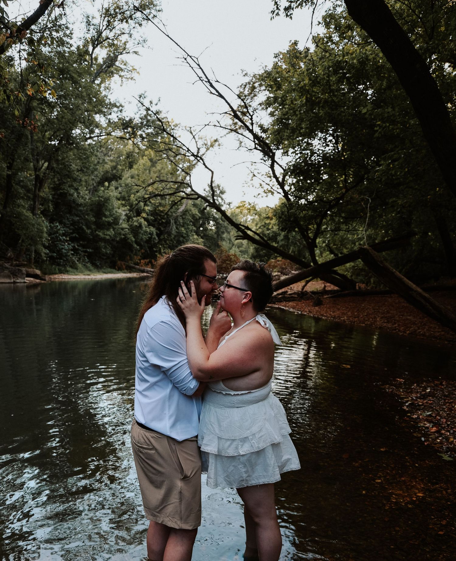 Couple sharing a romantic kiss while standing in a shallow river surrounded by lush green trees.