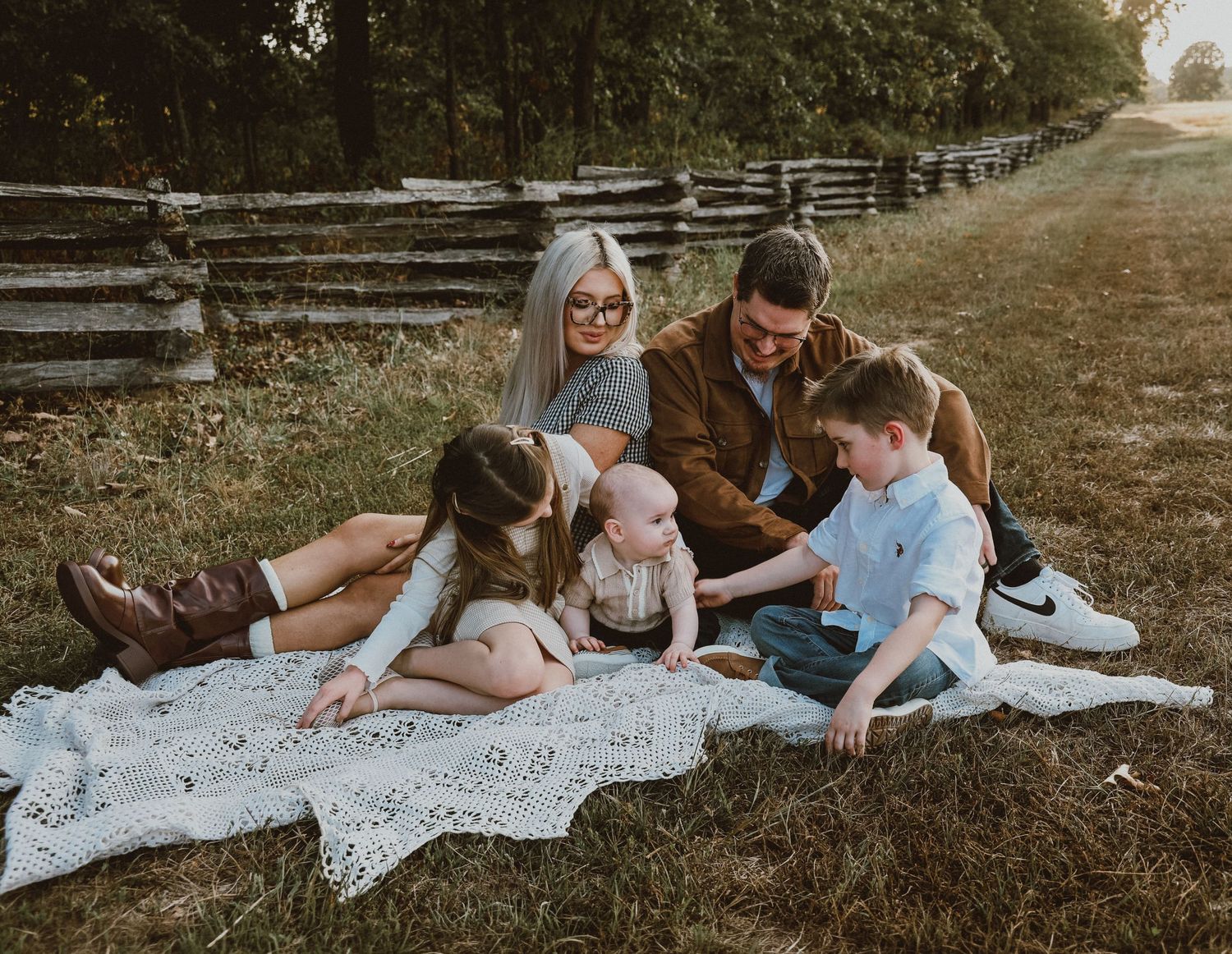 Family of five sitting on white blanket in grassy field near rustic wooden fence during outdoor photo session.