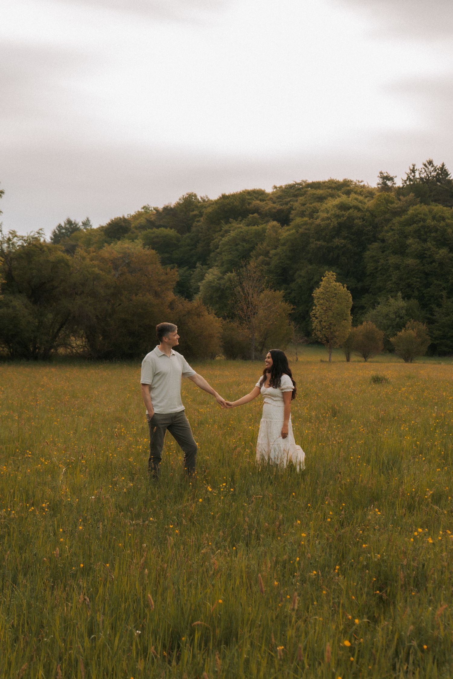 Couple holding hands in a lush green meadow with wildflowers, trees in background, woman in white dress.