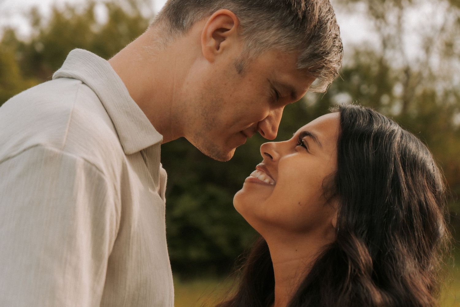 Romantic couple sharing an intimate moment outdoors, nearly kissing, with lush green trees in the background.