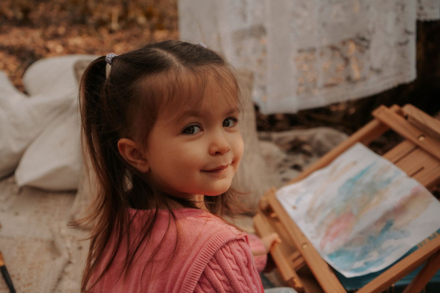 A smiling little girl with pigtails in a pink sweater sits outdoors near a colorful watercolor painting on an easel.