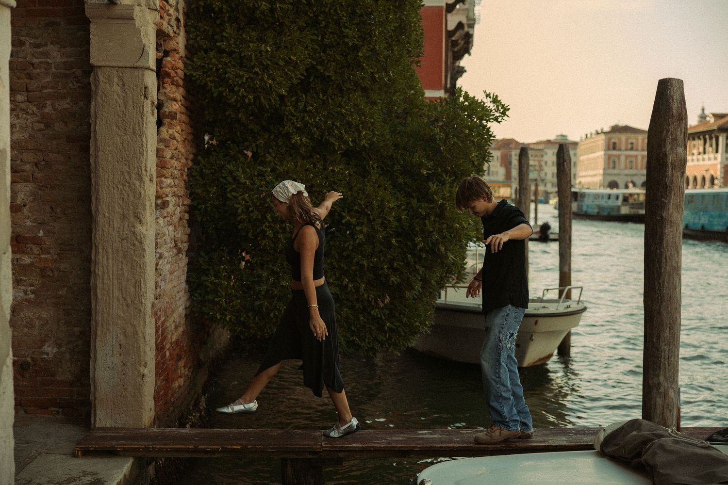 Two people balance on a narrow wooden plank crossing a Venice canal at sunset, with a moored boat and historic brick buildings nearby.