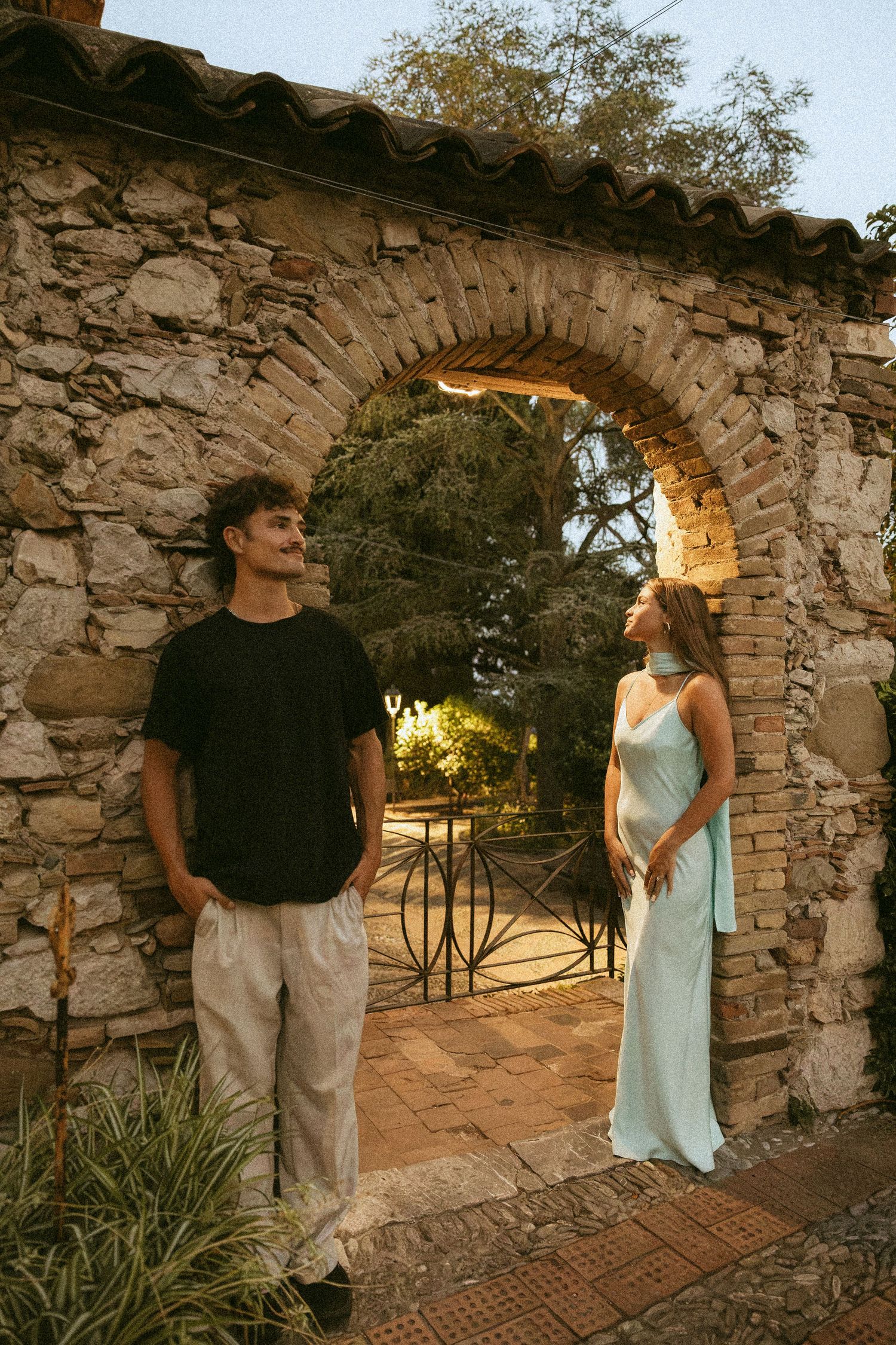 Man in black t-shirt and woman in a pale blue satin dress stand facing each other beneath a rustic stone archway in a sunlit garden.