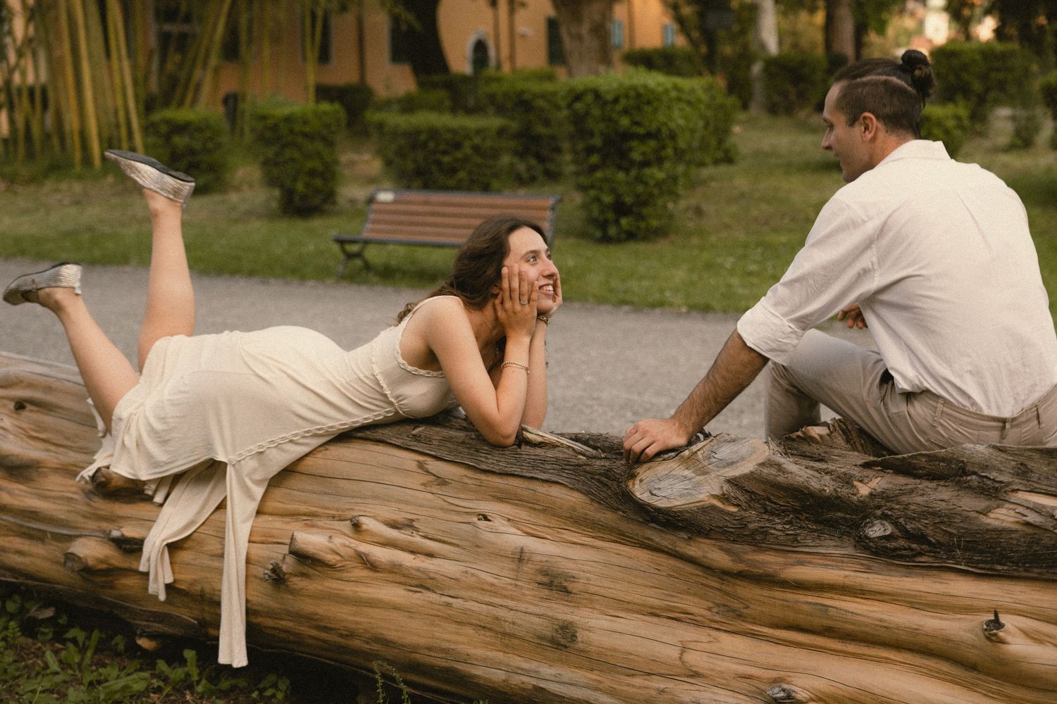 Woman in a white dress lying on a large fallen log smiling while a man in a light shirt sits beside her in a sunny park.