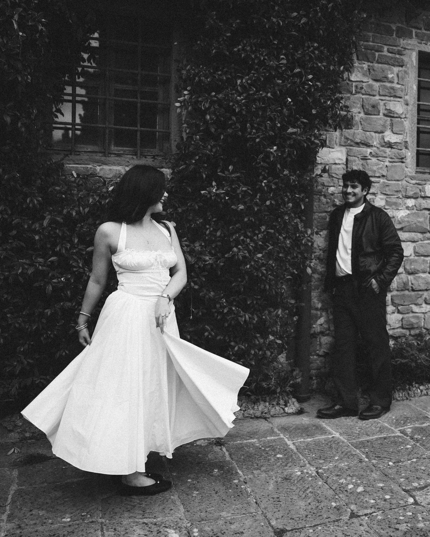 Black and white photo of a woman twirling in a flowing white dress as a smiling man leans against an ivy-covered stone wall.