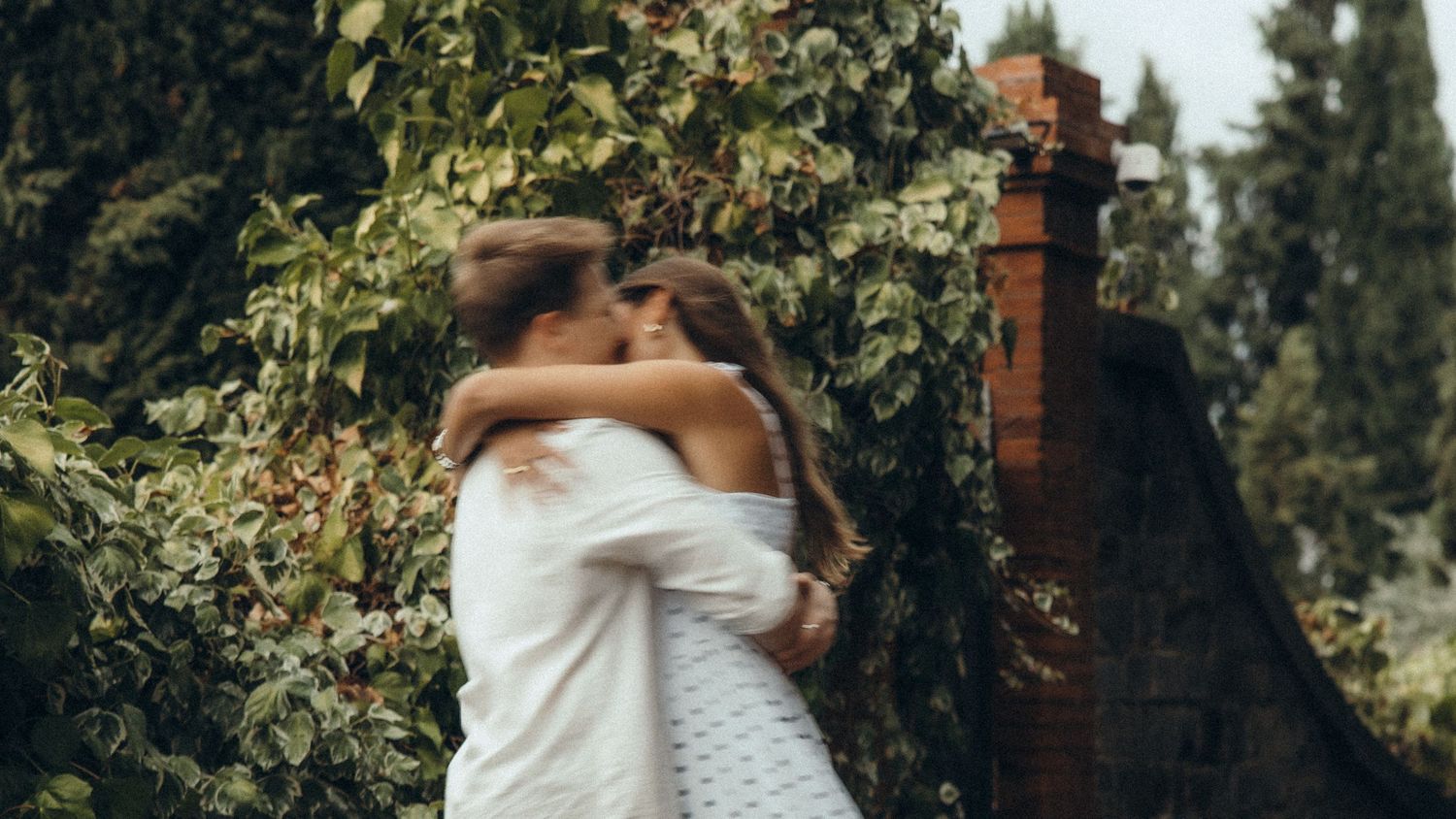 Couple in white outfits embracing and kissing outdoors beside a brick pillar covered in lush green ivy in a romantic garden setting.