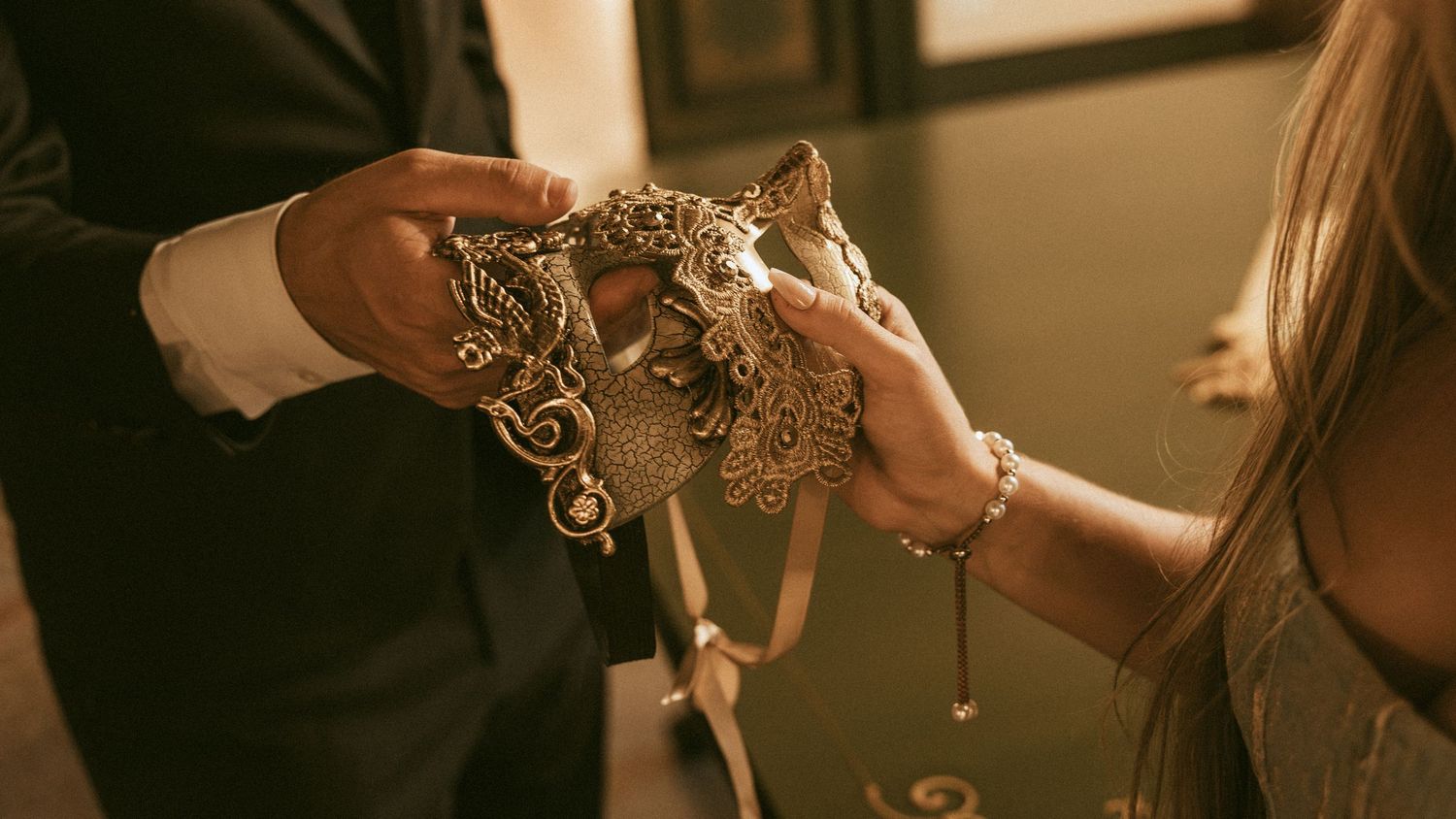 Man in a dark suit and a woman in a silver dress hold an ornate gold masquerade mask with ribbon ties, in warm light.