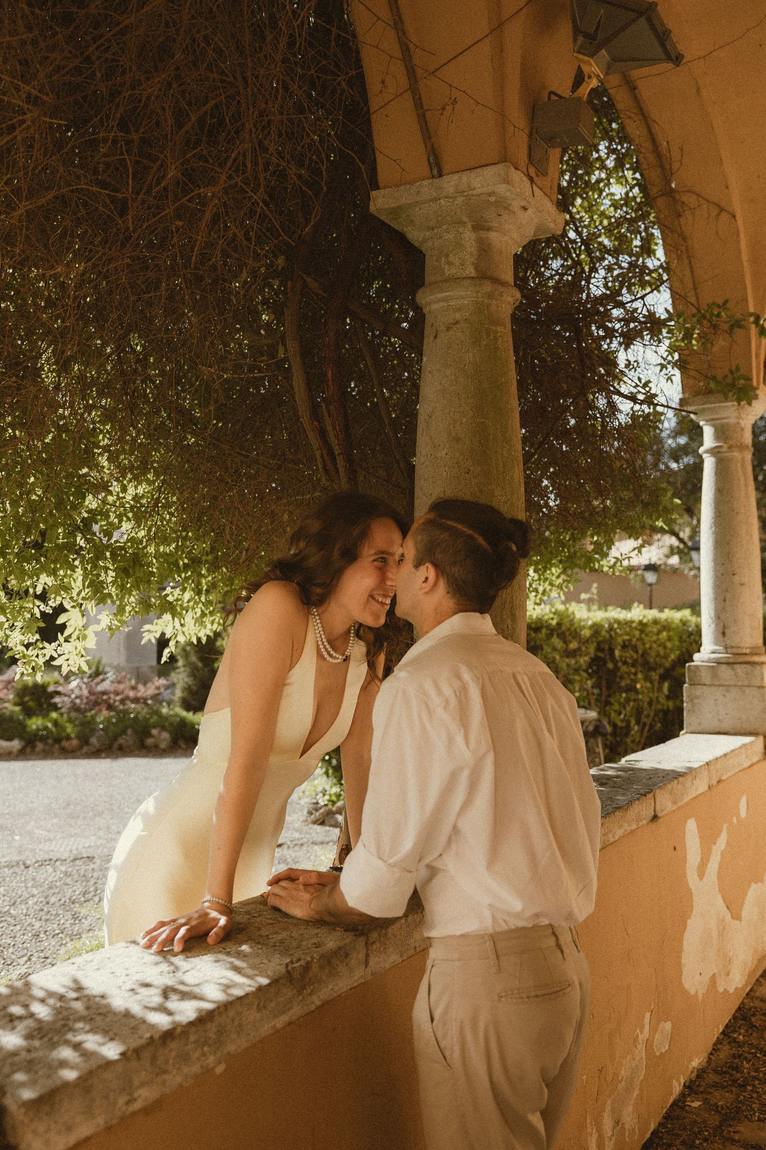 Romantic couple in a cream dress and white shirt share an intimate moment beneath a sunlit stone archway with climbing vines.