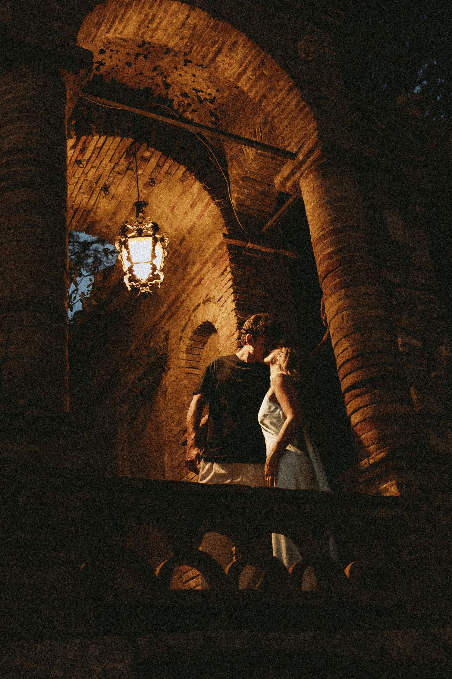 Couple embracing and about to kiss on a historic brick balcony beneath a glowing vintage lantern at night, in a romantic wedding portrait.