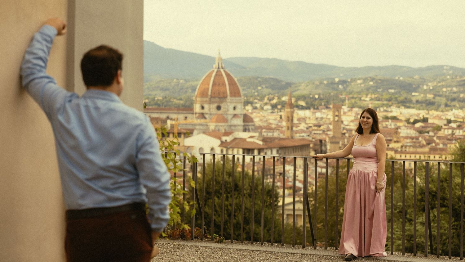 A man leans on a wall as a smiling woman in a pink dress stands at a railing with a Florence skyline and domed cathedral behind her.